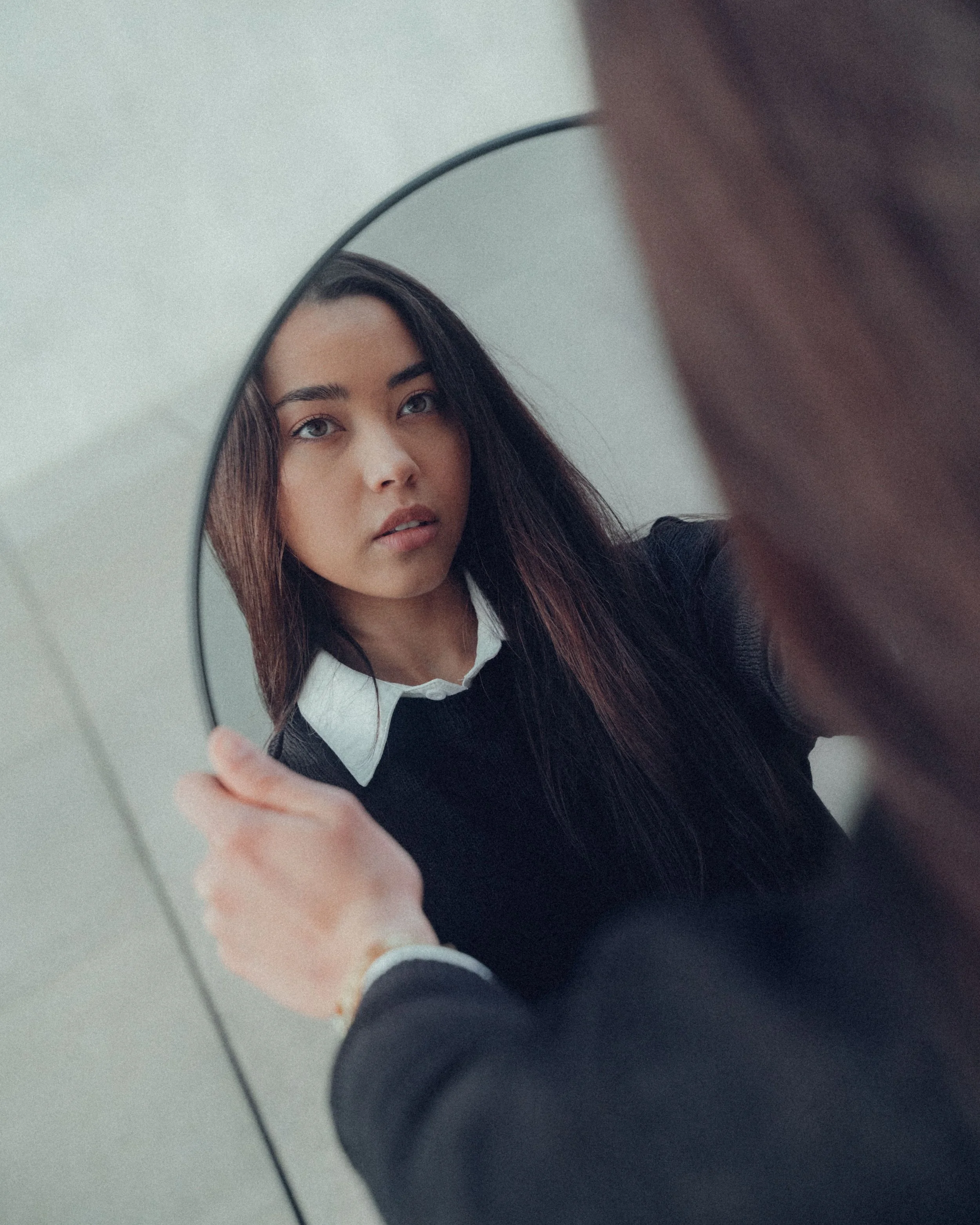 A young woman in a black sweater looks at her reflection in a round handheld mirror against a neutral background.