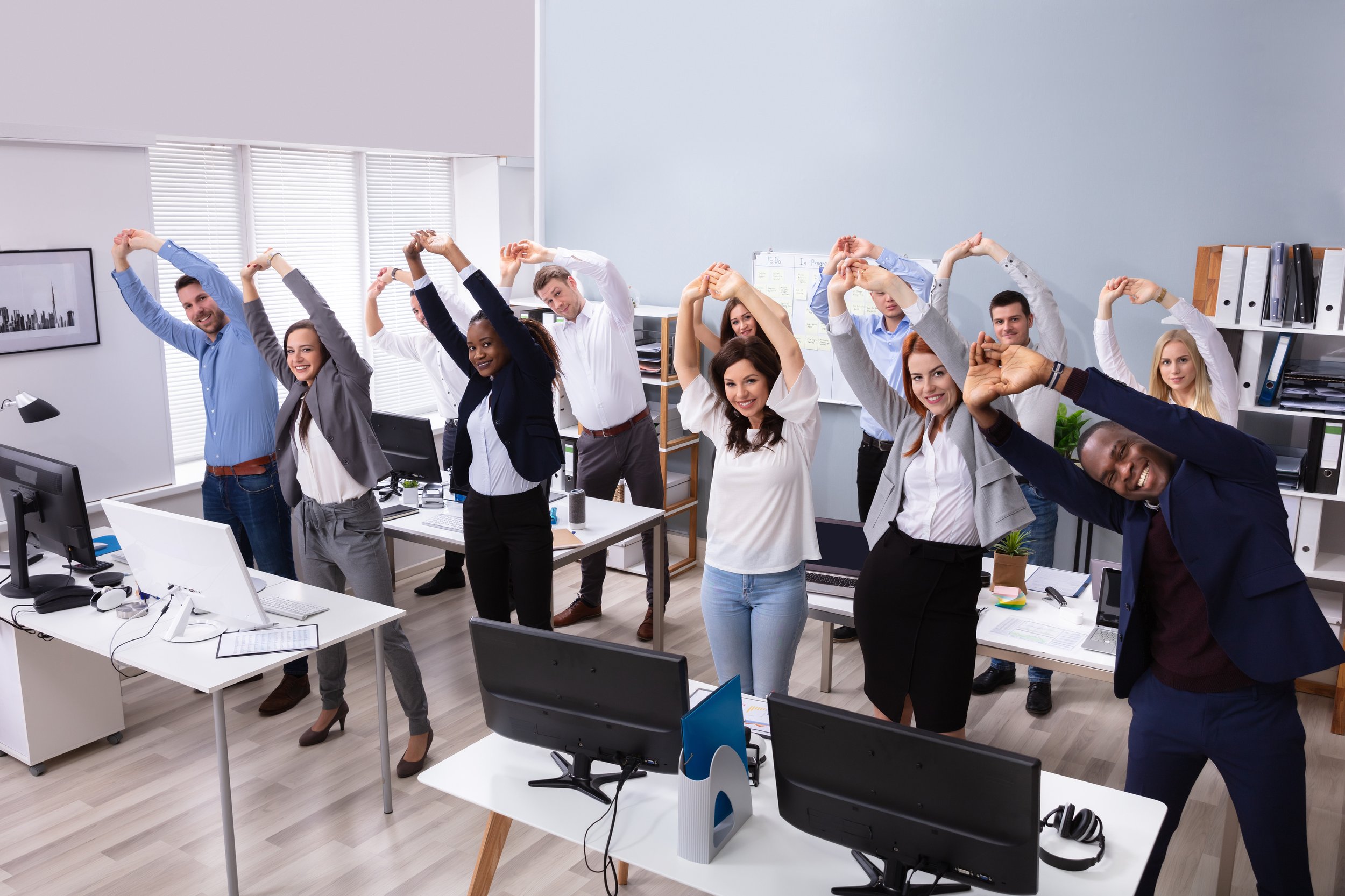  Group Of Smiling Multi-ethnic Businesspeople Doing Stretching Exercise At Workplace In Business Casual As Example Of Dougfit Corporate Health Events That Can Be Done In Office and In Business Attire  