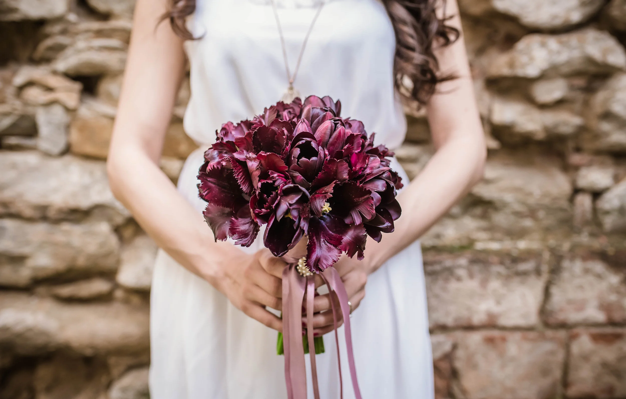A woman in a white dress holding a bouquet of dark purple flowers, standing in front of a stone wall.