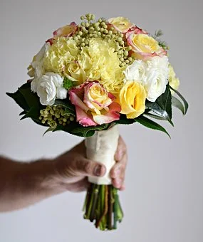 Hand holding a bouquet of yellow and pink roses, white carnations, and green foliage.