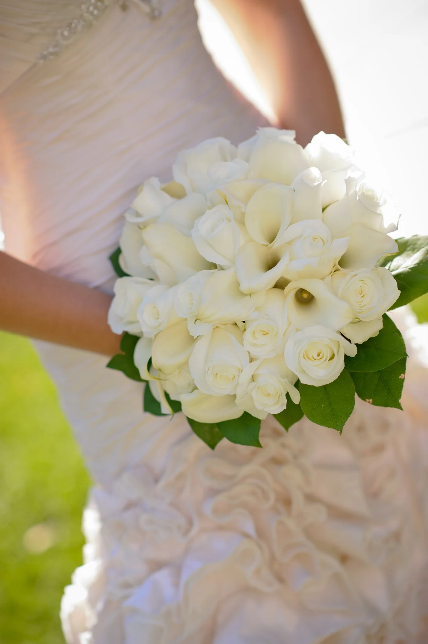 Close-up of a bride holding a bouquet of white roses and calla lilies in a wedding dress with textured fabric.