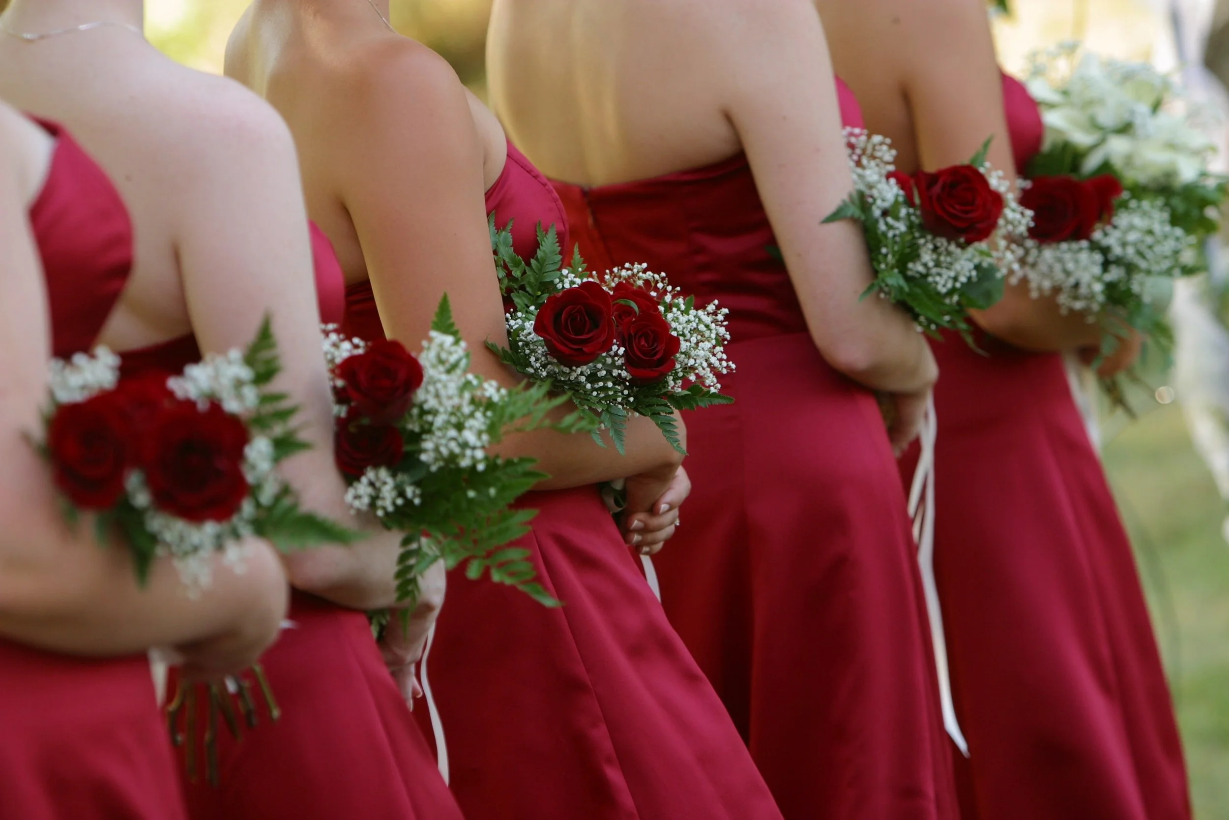 Bridesmaids in red dresses holding bouquets of red roses, white baby's breath, and green foliage at a wedding ceremony.