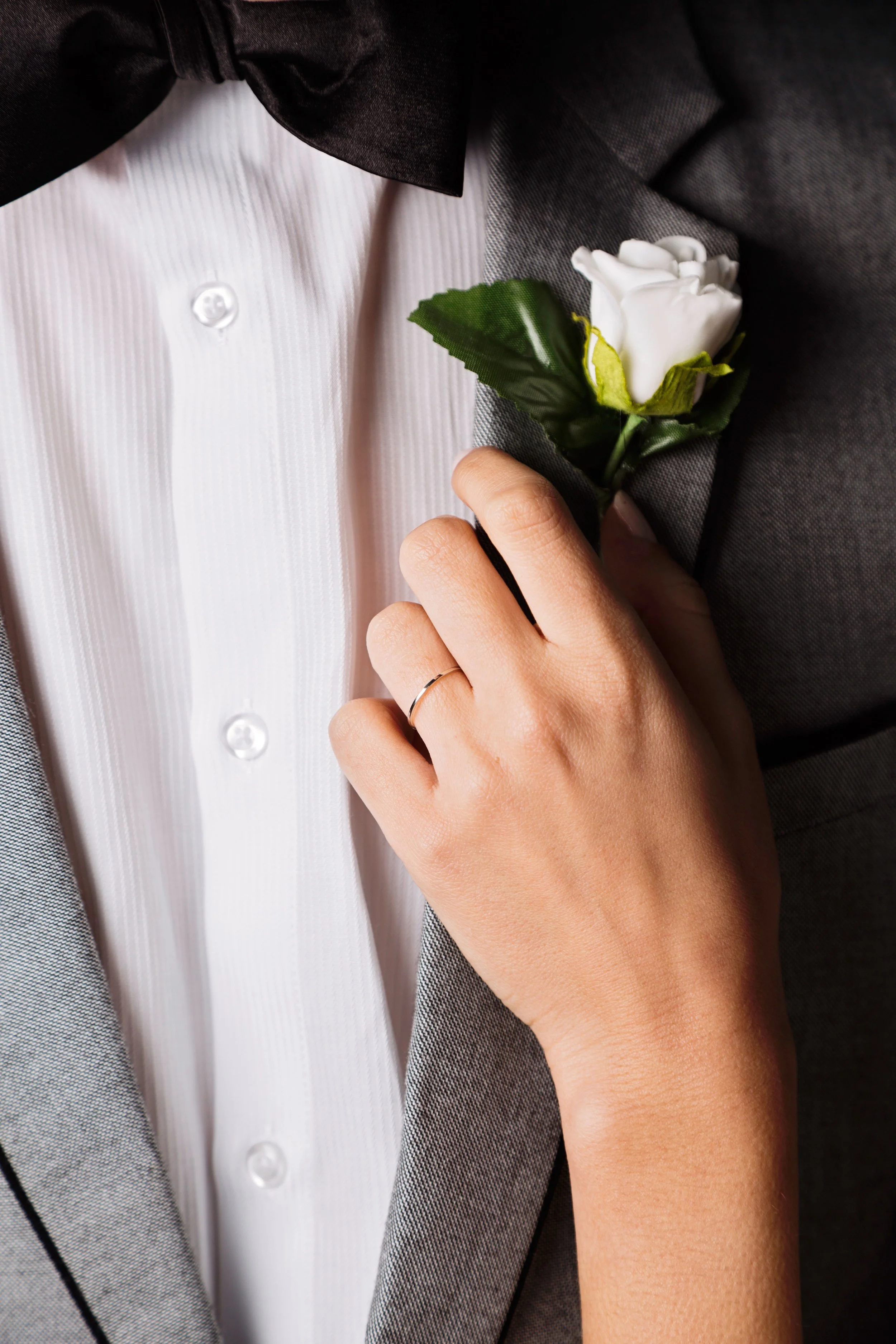 Close-up of a person in a tuxedo with a white dress shirt and black bow tie, placing a white flower boutonniere on the lapel.