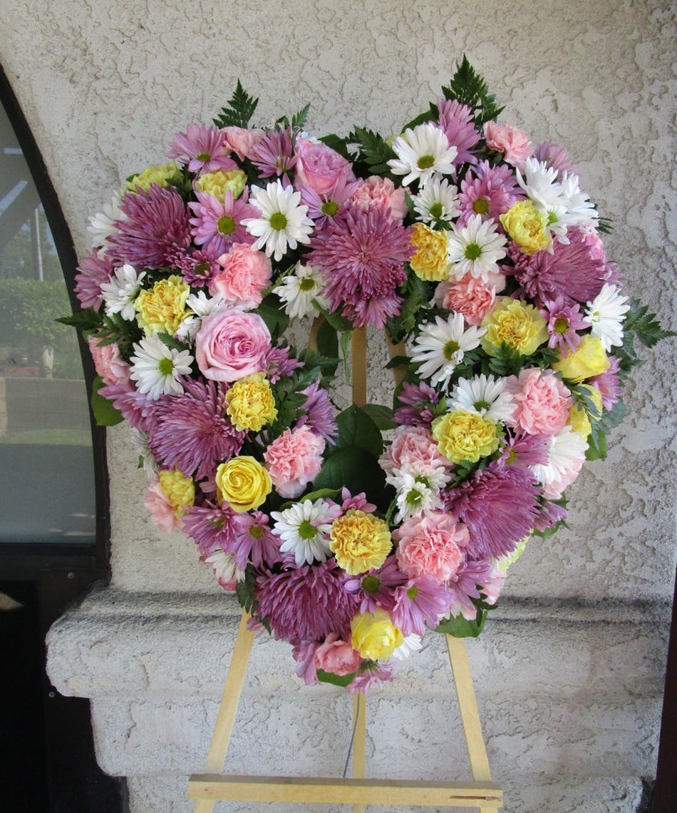 A large heart-shaped floral arrangement with pink, white, and yellow flowers on an easel outdoors against a beige textured wall.