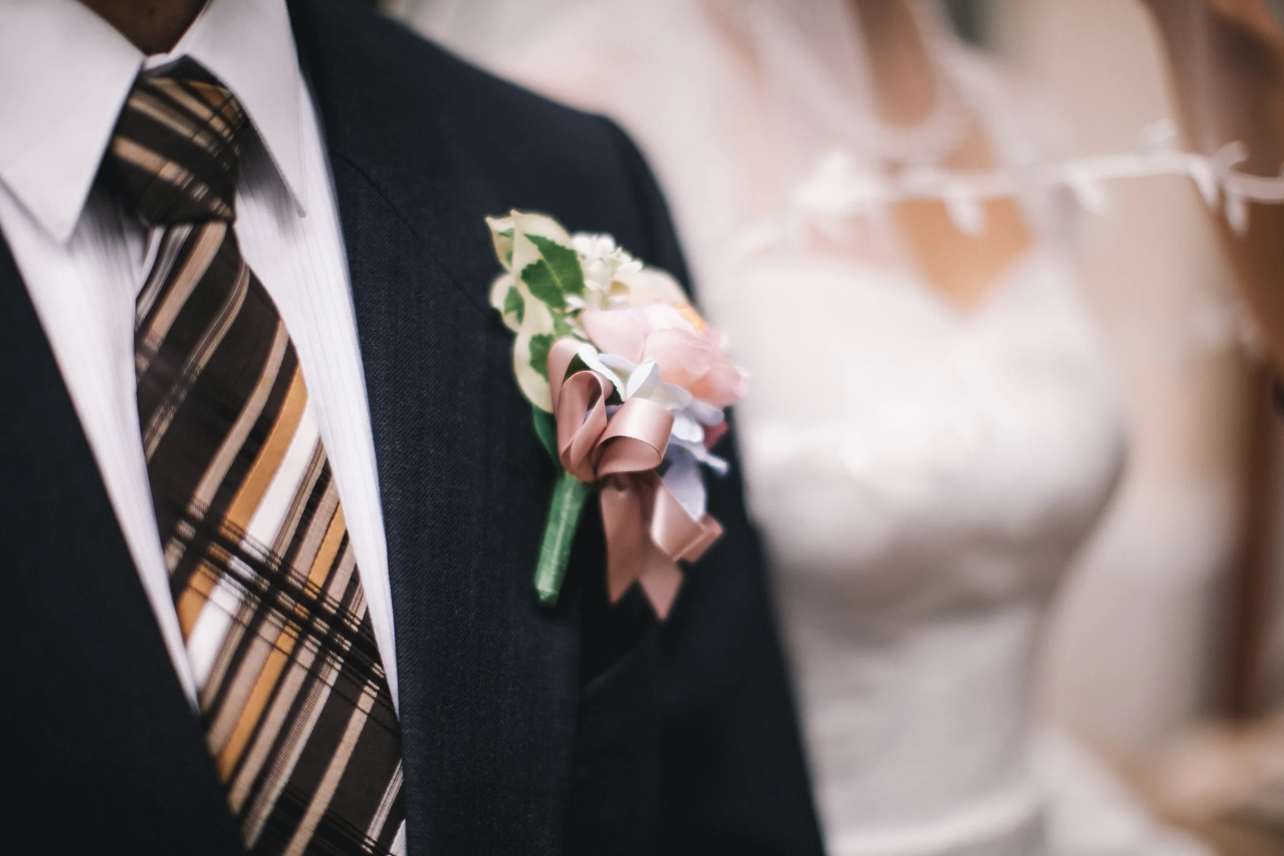 Close-up of a man in a dark suit with a plaid tie and a boutonniere of pink and white flowers with green leaves attached to his lapel, at a wedding ceremony.