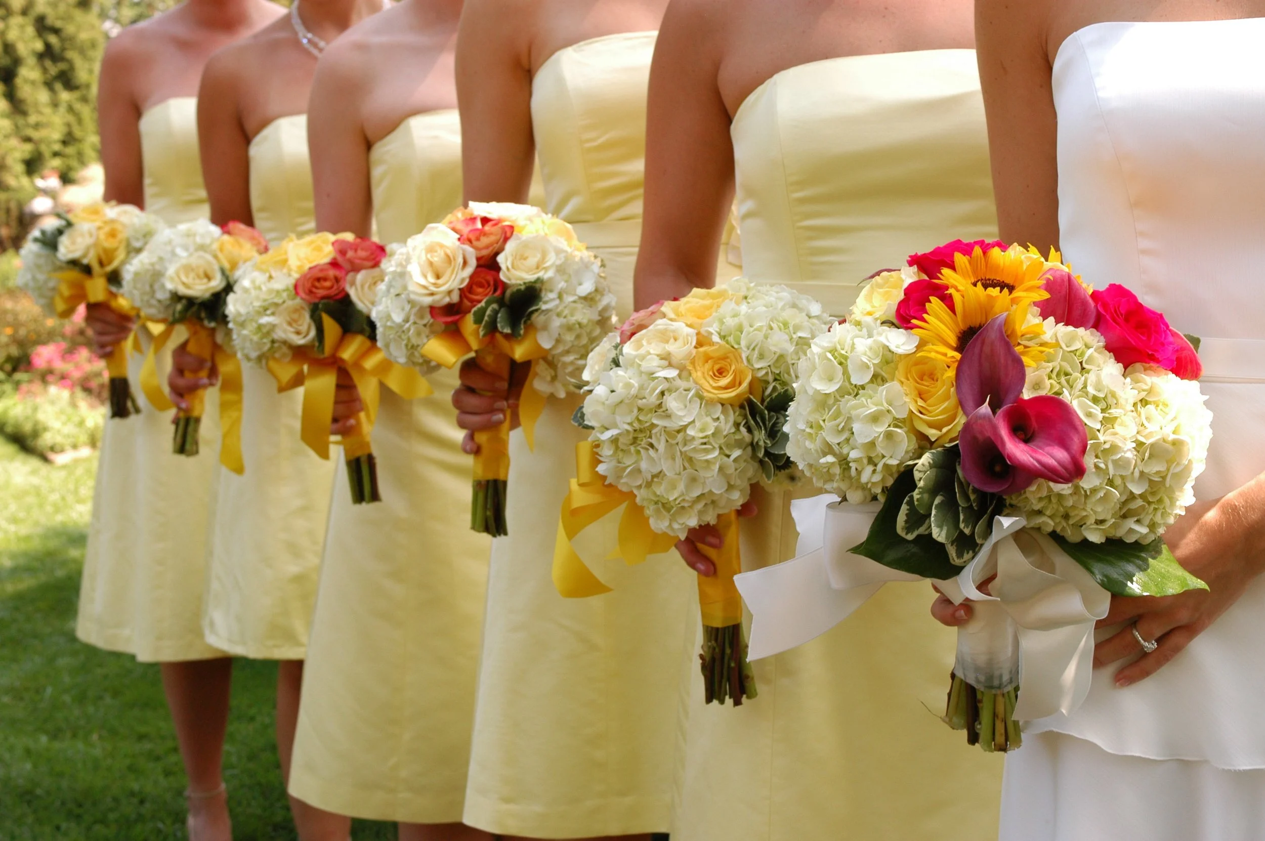 Bridesmaids in yellow dresses holding bouquets of white, yellow, and pink flowers at a wedding outdoors.