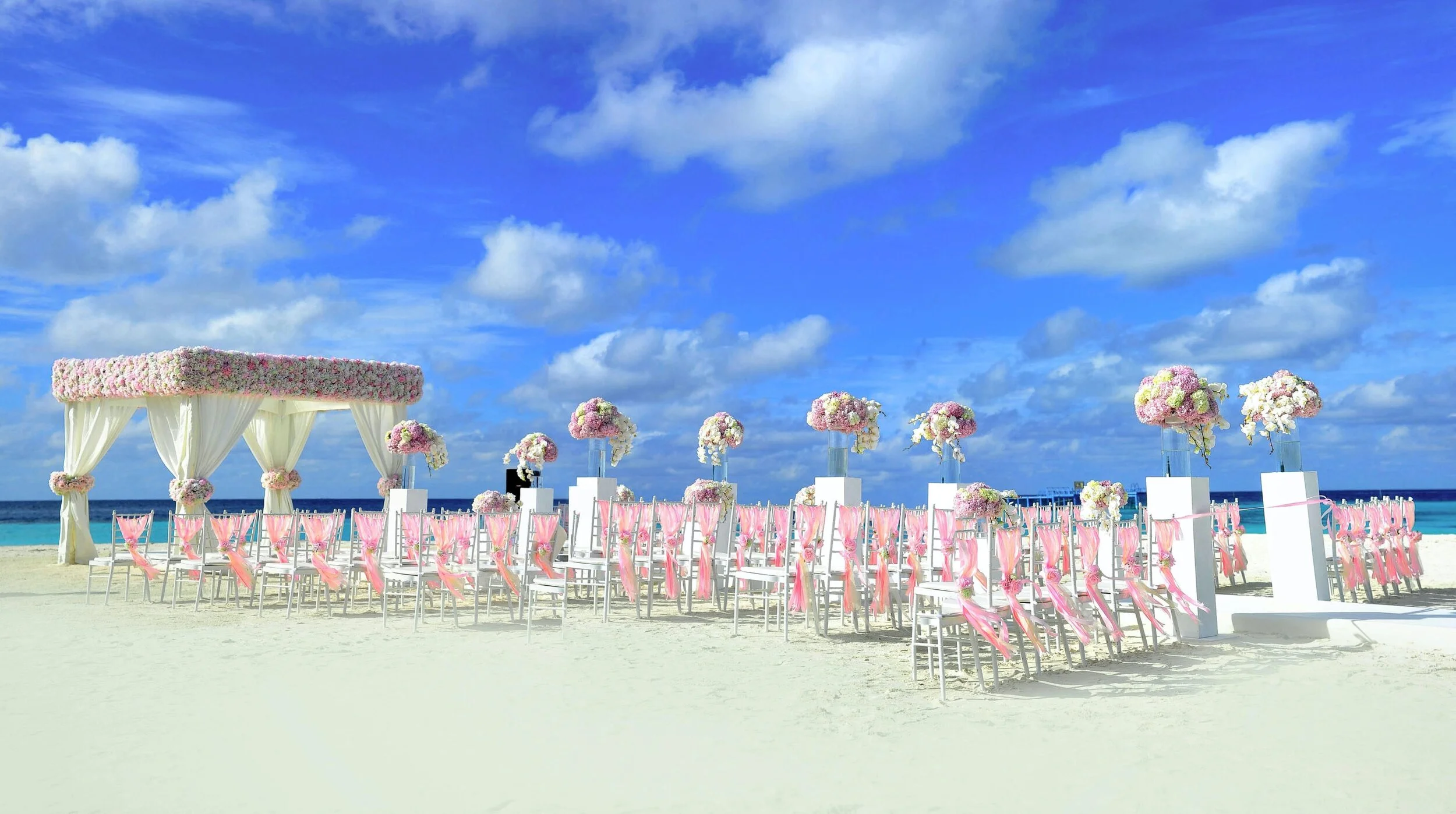 Decorated wedding ceremony setup on a beach with white chairs, pink ribbons, flower arrangements, and a flower-covered canopy under a blue sky with scattered clouds.