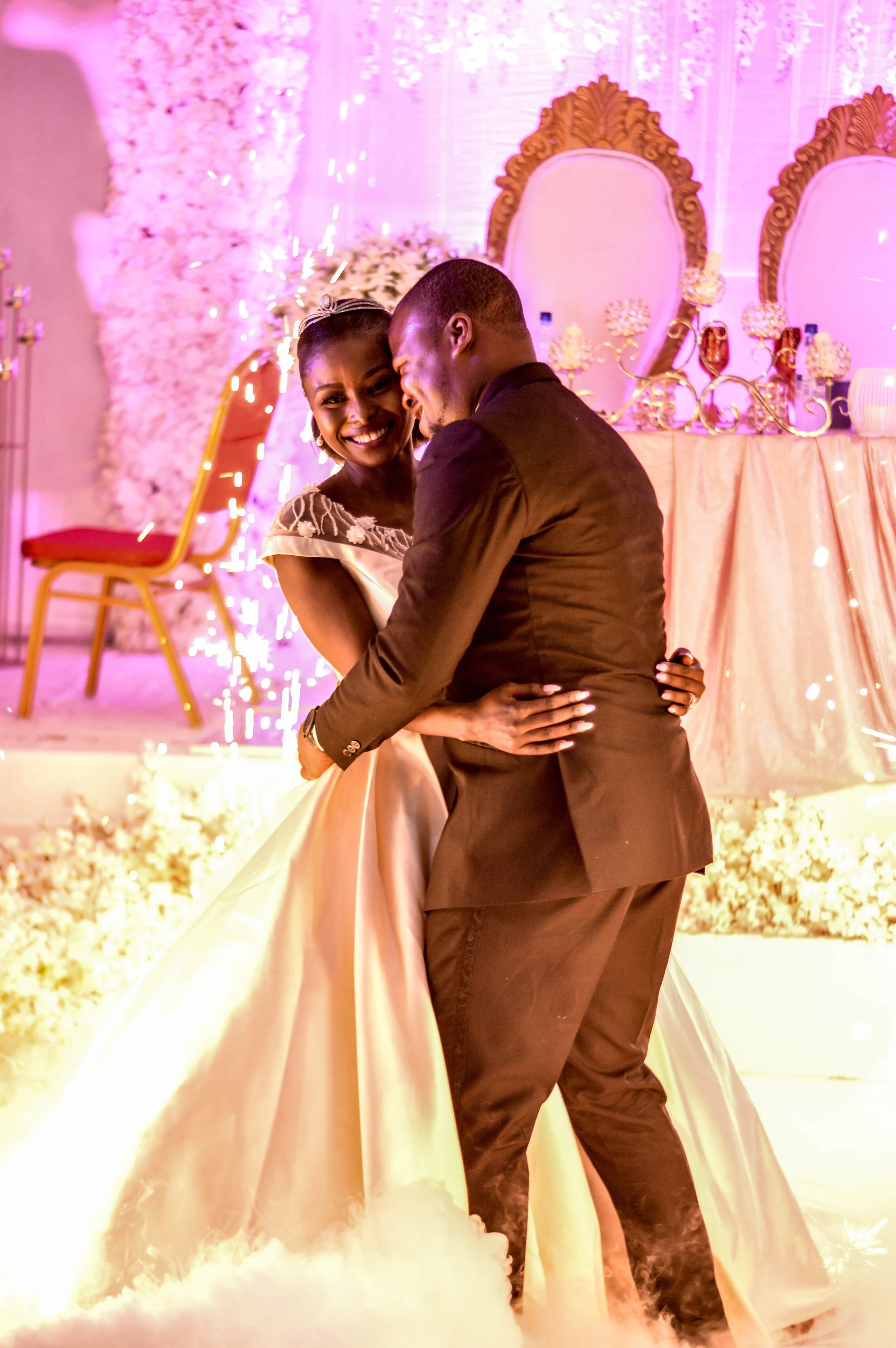A bride and groom dancing at their wedding reception, smiling and embracing each other with a pink and white decorated background, including floral arrangements and chairs.