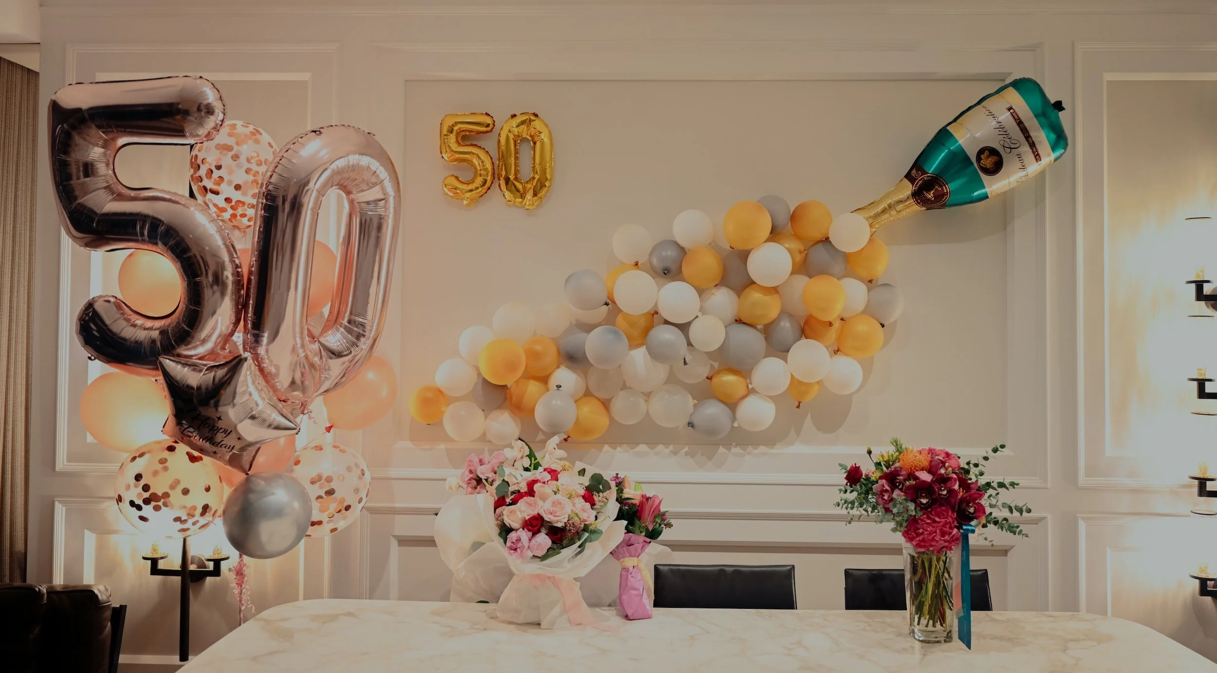 Decorated wall with balloons forming the number 50, a large balloon bouquet, a paper lantern, and balloons in white, gold, gray, and pink, along with floral arrangements on a dining table.
