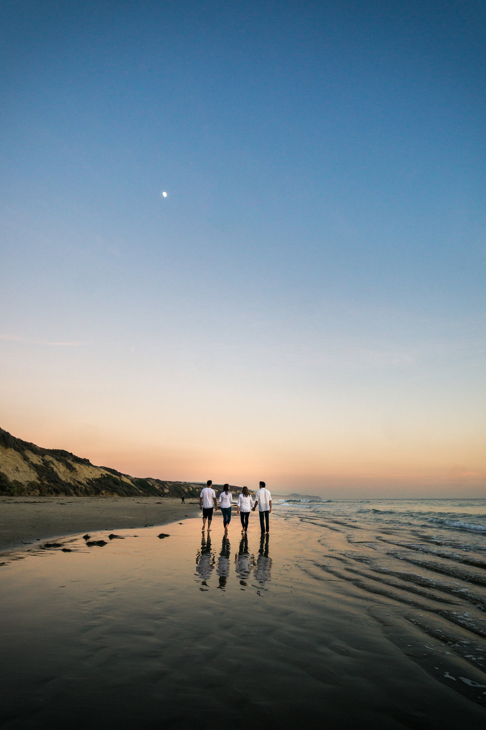 Family portraits of family walking on glassy seashore with blue sky and crescent moon while enjoying the sunset together during Golden hour at Crystal Cove State Beach in Newport