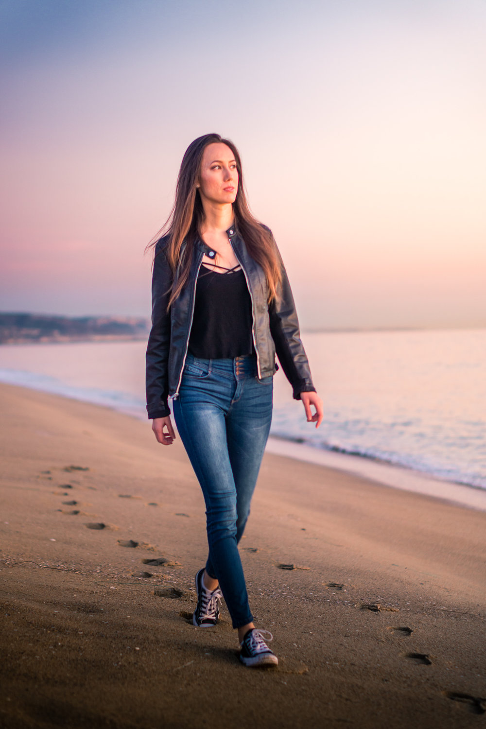 vibrant Natural light Fashion Portrait of woman wearing black leather jacket and blue jeans walking on beach during Golden hour At Balboa Pier in Orange County