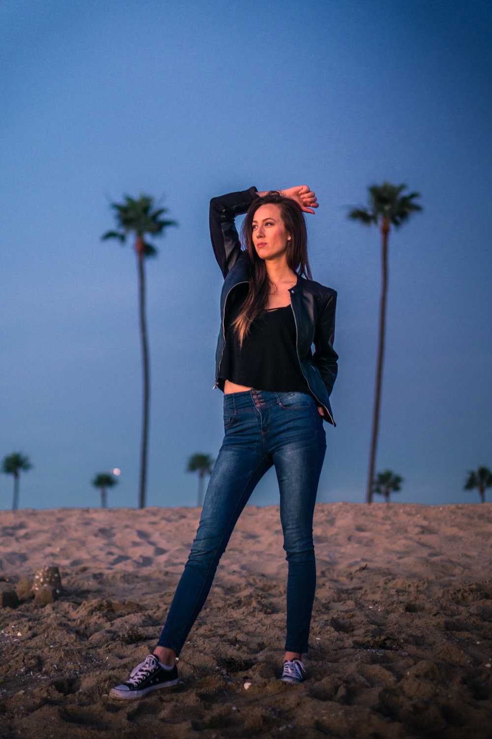 vibrant Natural light Fashion Portrait of woman wearing black leather jacket and blue jeans walking on beach during Golden hour At Balboa Pier in Newport Beach