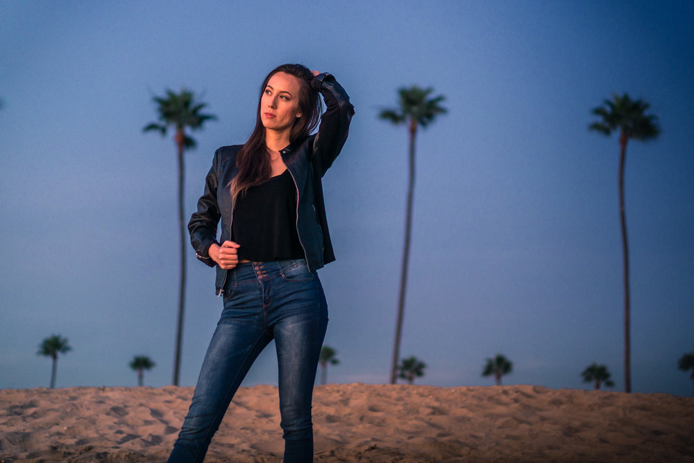 Natural light Fashion photoshoot of model posing in black leather jacket and blue jeans with blue sky and palm trees during Golden hour At Balboa Pier in Newport Beach