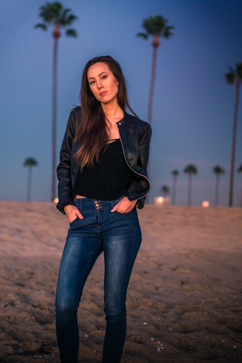 Natural light Fashion photoshoot of model posing in black leather jacket and blue jeans with blue sky and palm trees during Golden hour At Balboa Pier in Newport Beach