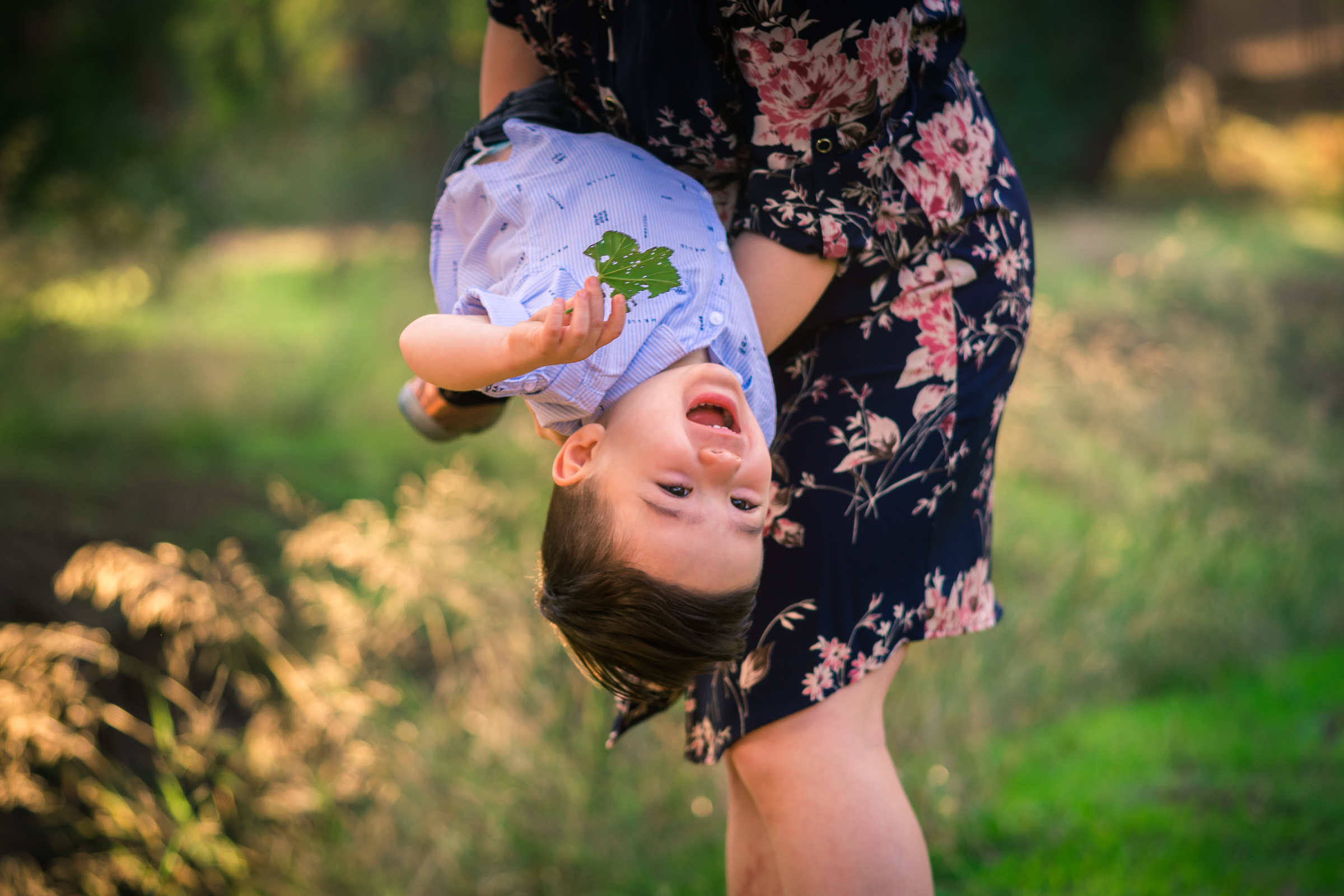 Candid photo of a little boy laughing with mother during a Family portrait photo shoot in orange county on the Juanita Cooke Trail with vibrant green trees and grass and the golden hour sun