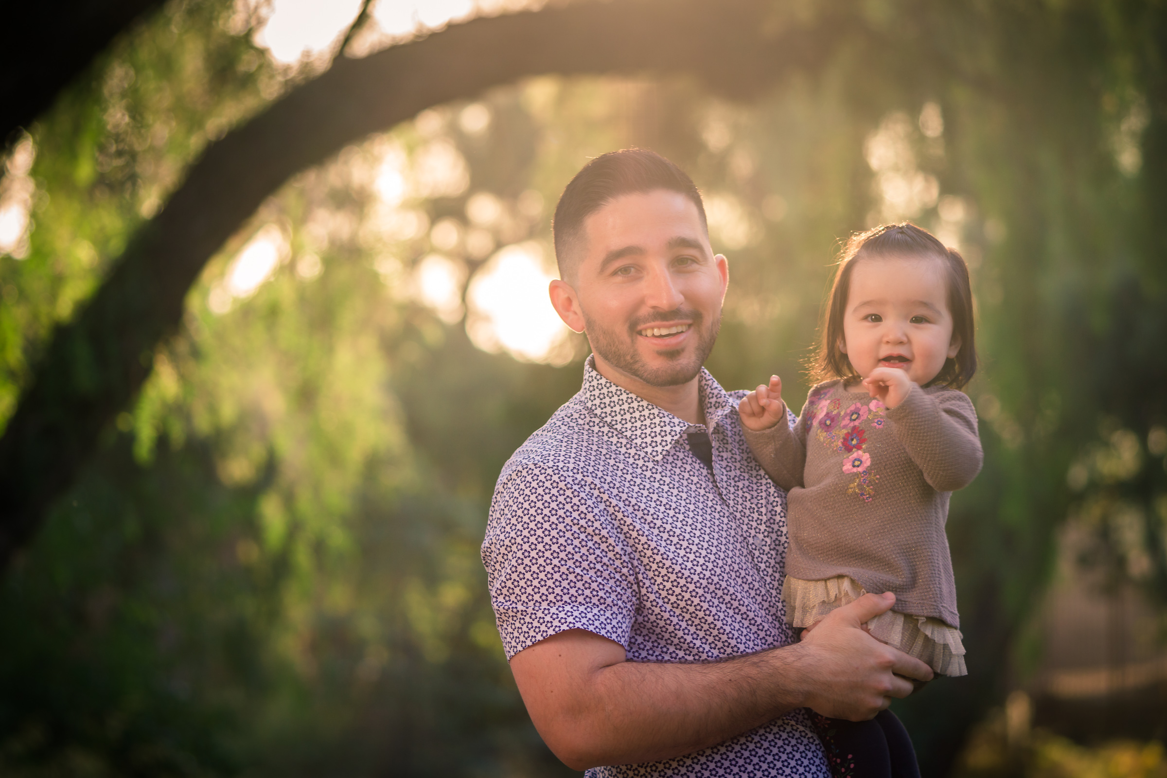 Candid photo of a little girl smiling with father during a Family portrait photo shoot in orange county on the Juanita Cooke Trail with vibrant green trees and grass and the golden hour sun