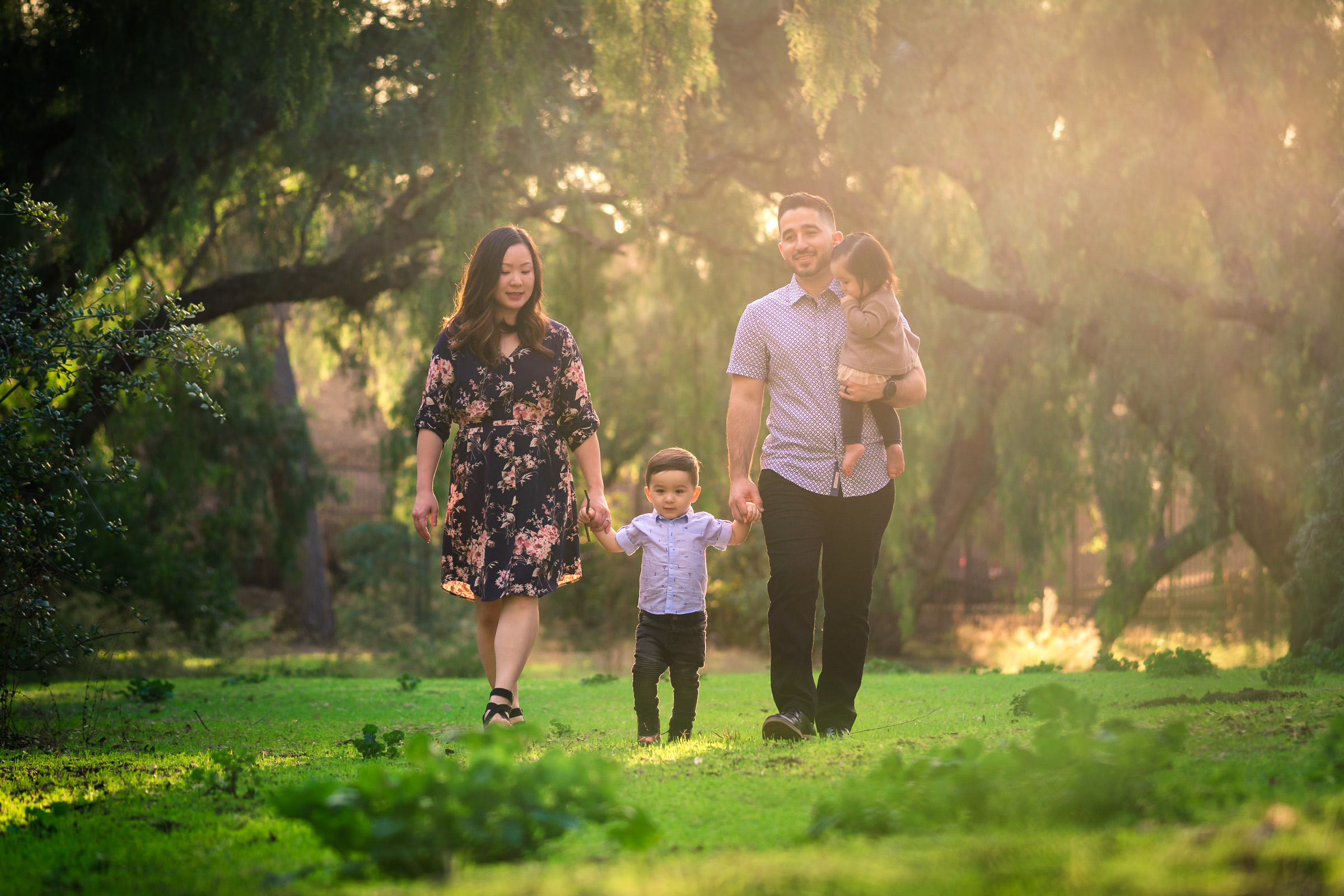 Candid photo of family walking during a Family portrait photo shoot in Fullerton on the Juanita Cooke Trail with vibrant green trees and grass and the golden hour sun