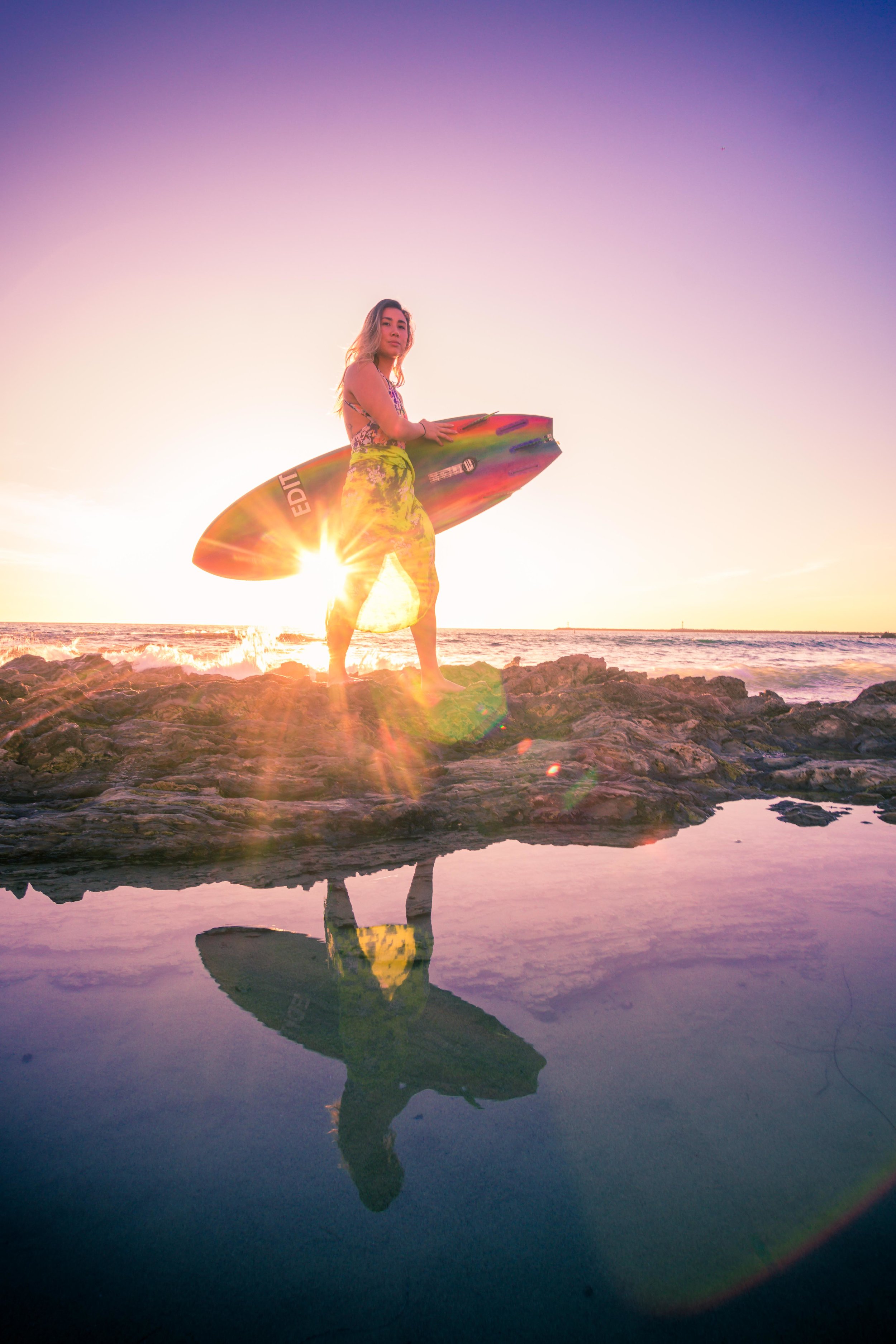 Surfer girl walking across the tide pools at little corona beach with her surfboard during a vibrant sunset