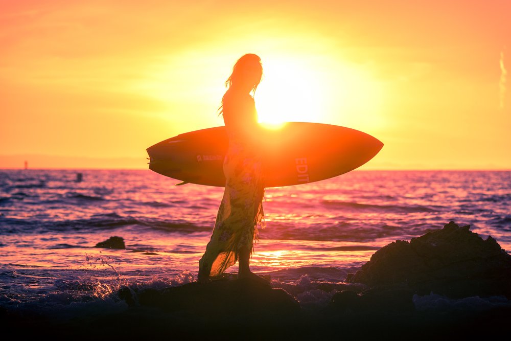 Hey beautiful artistic surfer girl portrait of her silhouette while she is holding her surfboard at the little beach tidepools in Orange County with an orange and purple sunset