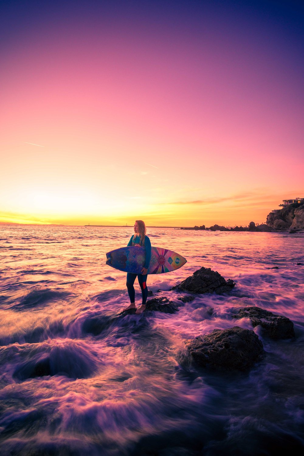 A wide-angle portrait of a surfer girl standing in the waves using long exposure techniques during a Golden and purple California sunset at little corona beach in orange county