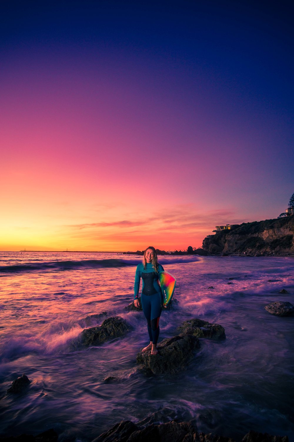 A portrait of a surfer girl at little corona beach with her standing on the tidepool rocks with the waves breaking during a beautiful purple sunset