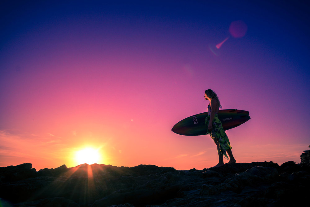 a silhouette of a surfer girl holding her surfboard on the rocks at corona Del mar beach at sunset
