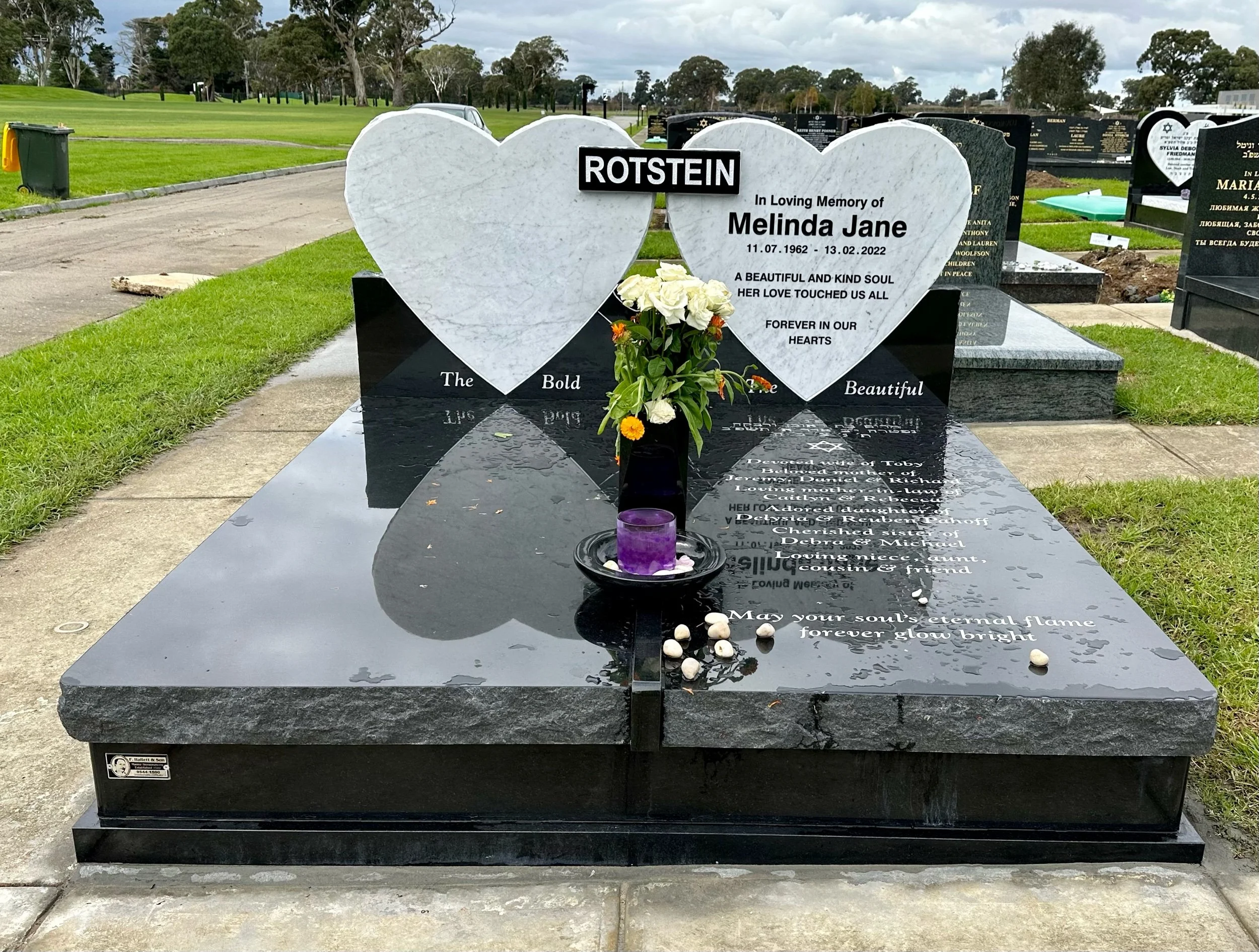 A grave marker at a cemetery with two large white heart-shaped stones. The left heart has the name 'ROTSTEIN' and the right heart has the inscription for Melinda Jane, showing her birth and death dates, and a memorial message. There is a black vase with white and yellow flowers, a purple candle, and white stones on the grave.