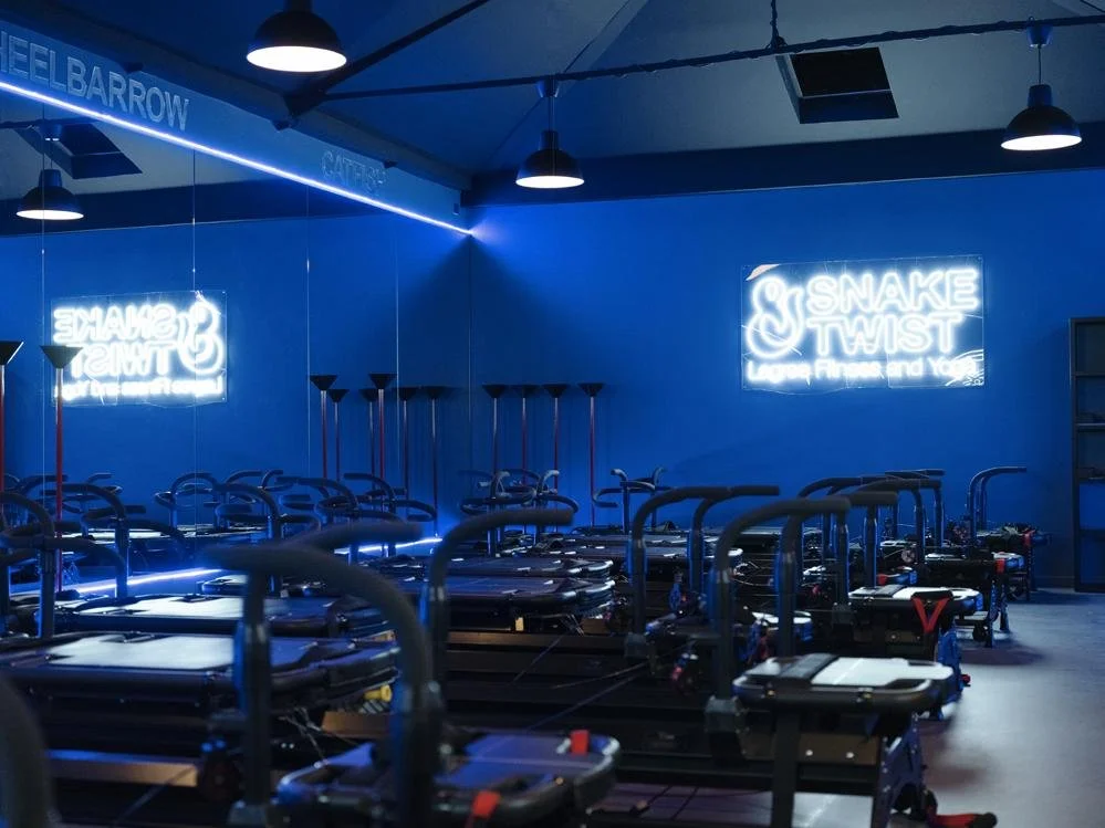 Indoor fitness studio with black reformer Pilates machines arranged in rows, illuminated by blue neon lights, with a mirror on the wall reflecting the equipment and neon signs that read 'SNAKE & TWIST' and 'SHAPE & STRENGTH'.