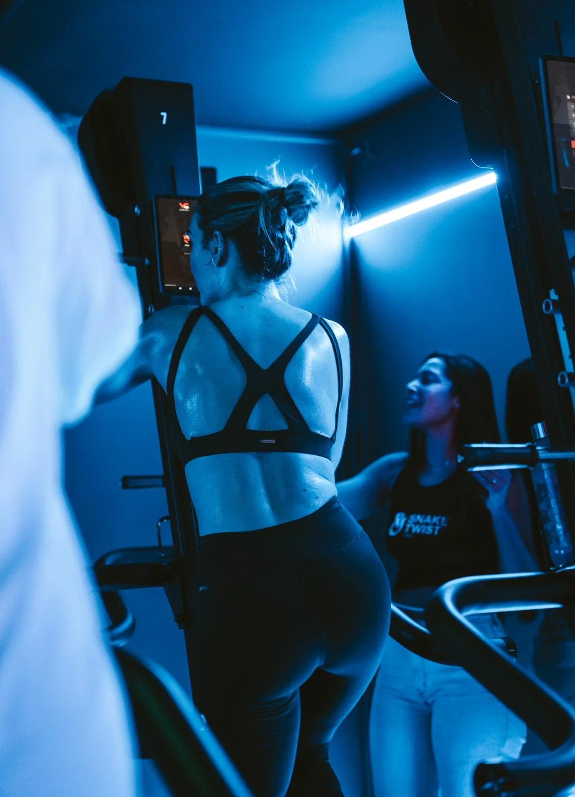 Two women at a gym; one woman in a black sports bra and leggings is exercising on a machine, while another woman in a black shirt and white pants is standing nearby talking.