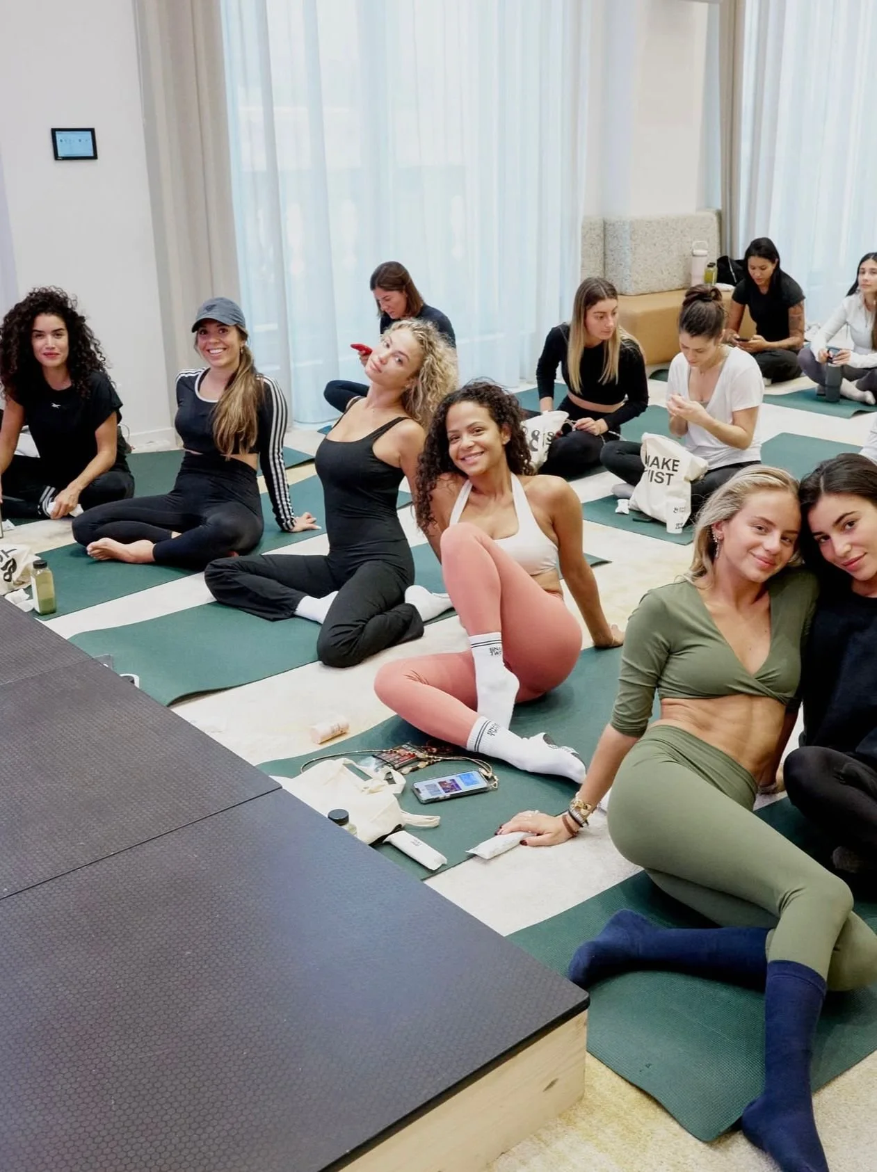 Group of women participating in a yoga or fitness class in a bright room with large windows and light curtains, sitting or stretching on mats with personal belongings nearby.
