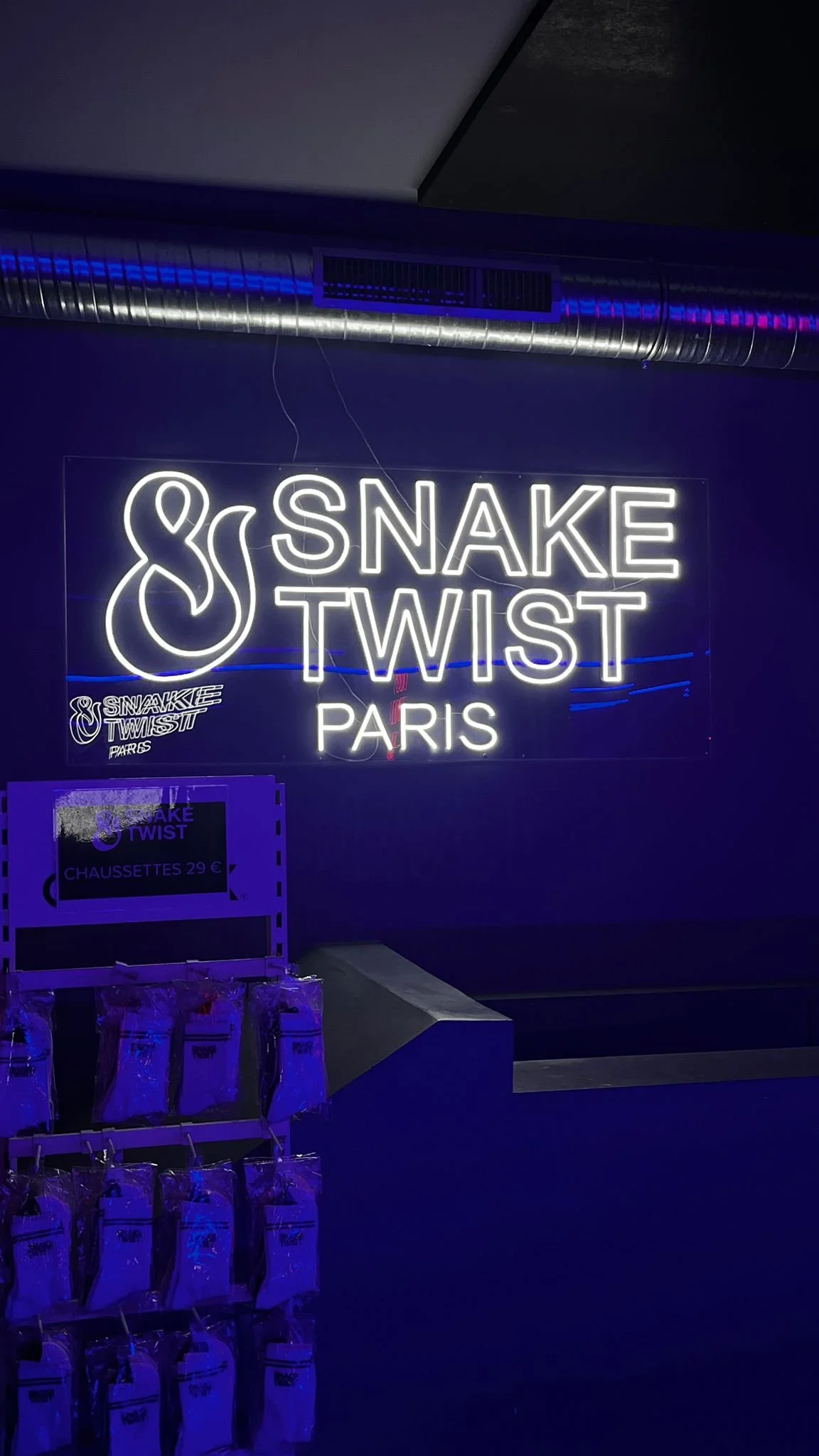 Neon sign displaying the text 'Snake Twist Paris' with a graphic of a snake, located indoors with a dark wall and metallic air duct above.