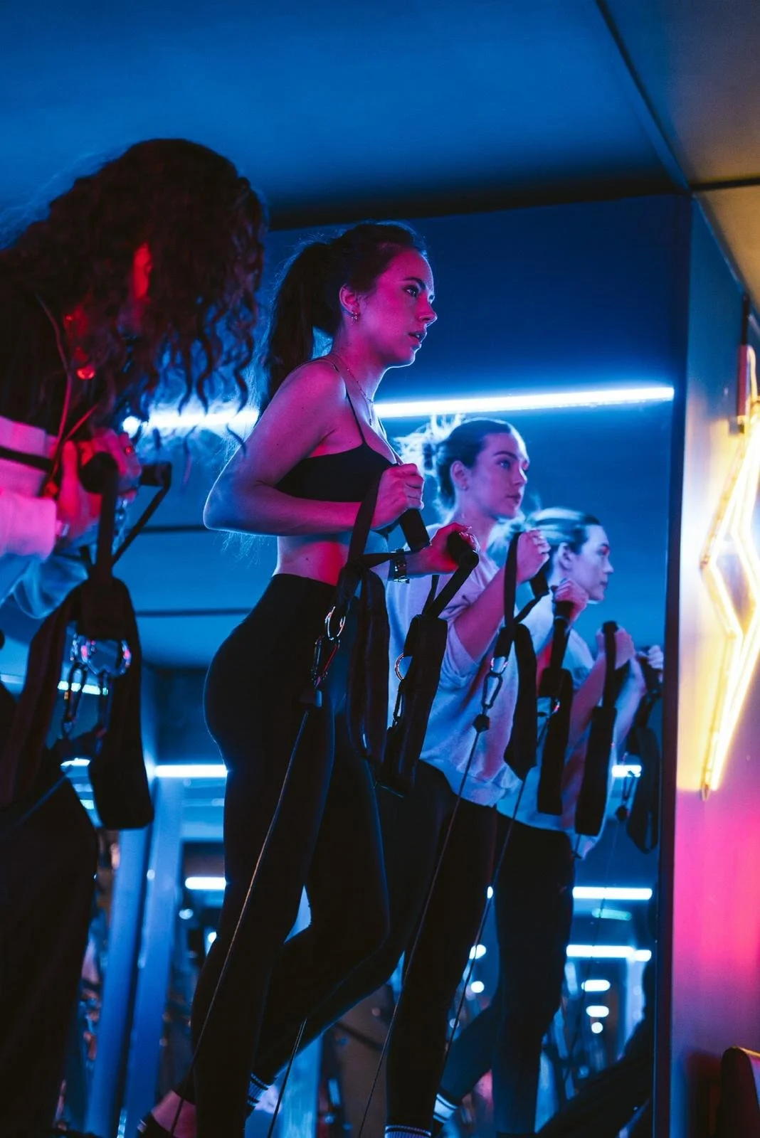 Four women exercising with resistance bands in a fitness class, illuminated by blue and neon lights.