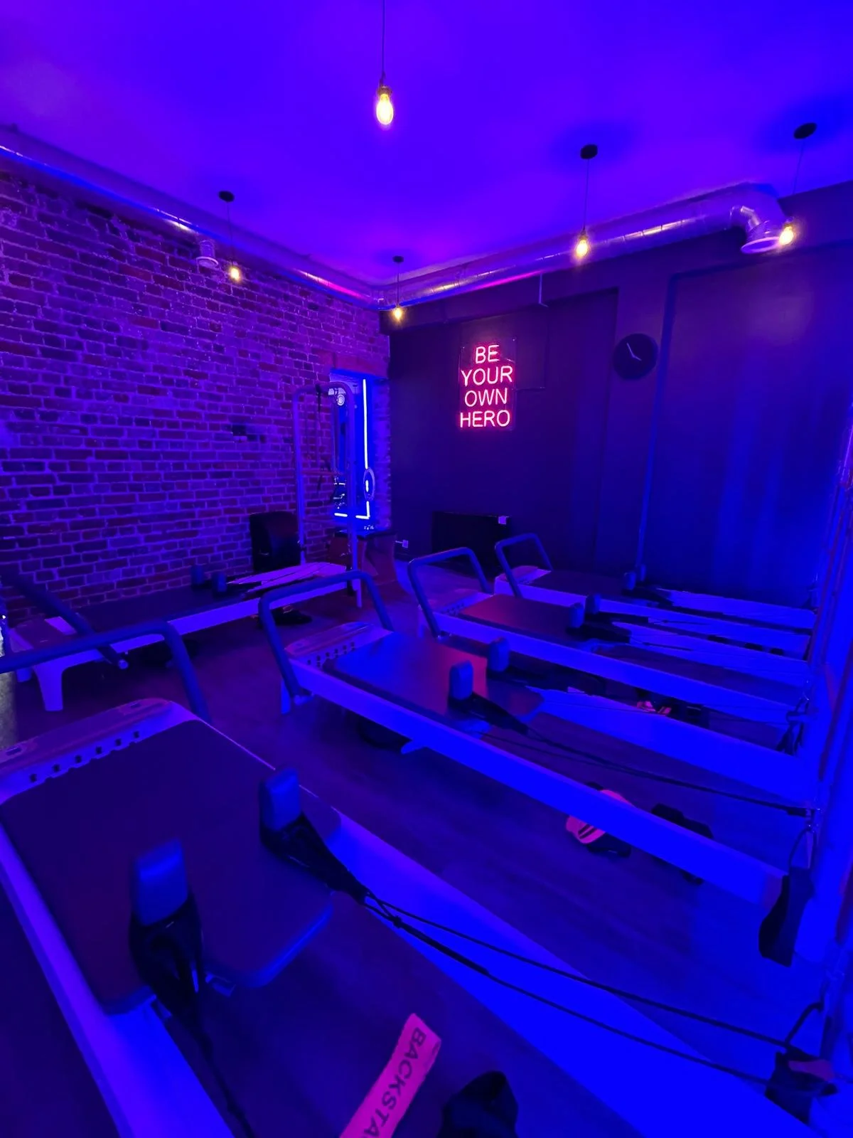 Room with multiple rowing machine exercise equipment, illuminated by purple and blue lighting, brick wall with neon sign that reads 'BE YOUR OWN HERO', and exposed ceiling pipes.