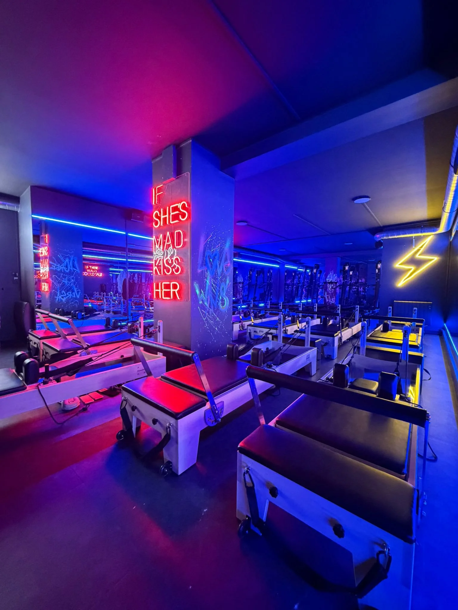 Fitness studio with Pilates reformer machines, colorful neon lights, a wall with graffiti and neon signs, including one that says 'If she’s mad kiss her'.