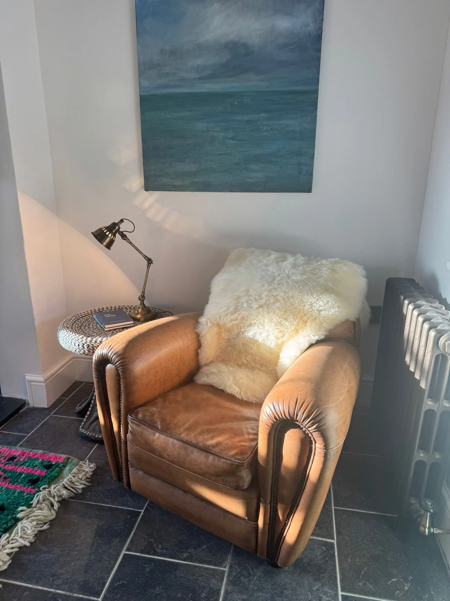 A cozy living corner with a brown leather armchair topped with a fluffy cream-colored pillow, next to a black radiator and a small round side table with a black and white woven pattern holding a small notebook and a brass desk lamp, with a large abstract painting of a blue ocean scene on the white wall behind.
