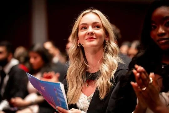 A young woman with long blonde hair sitting in a conference or seminar room, holding a colorful brochure. She is smiling and looking slightly to the side, with other attendees blurred in the background.