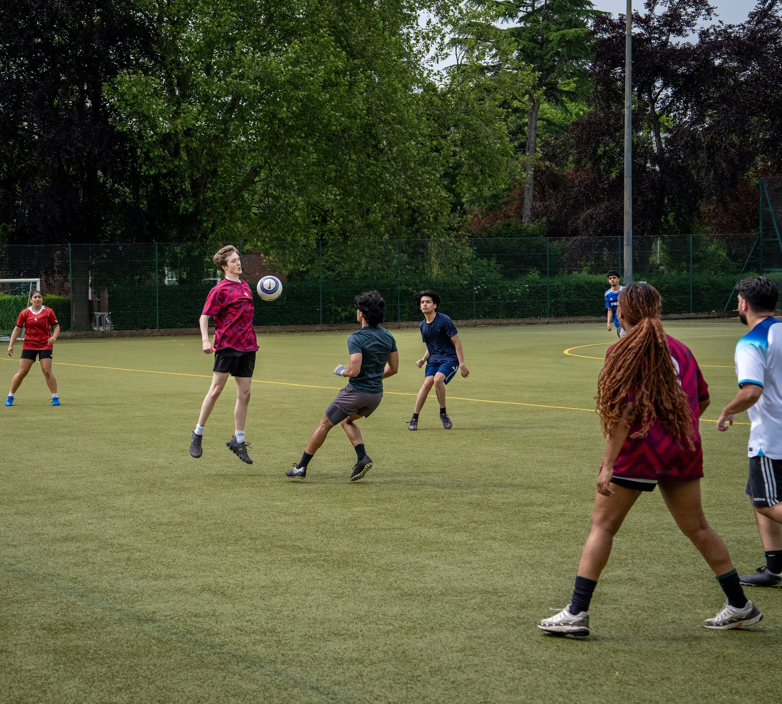 People playing football on a field surrounded by trees and a fence.