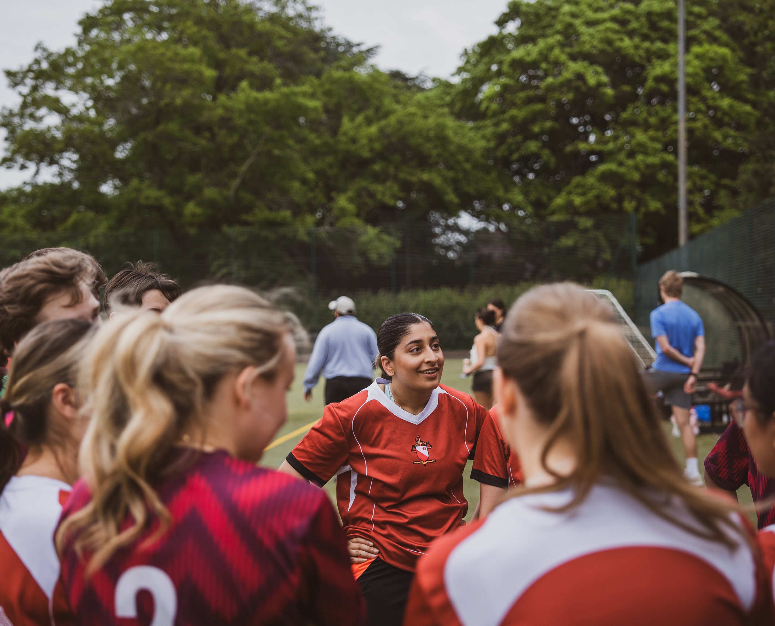 A group of people gathered outdoors on a sports field, with a Jasmine Khak in a Leicester University Law Society's sports jersey in the center, smiling and talking to others, surrounded by trees.