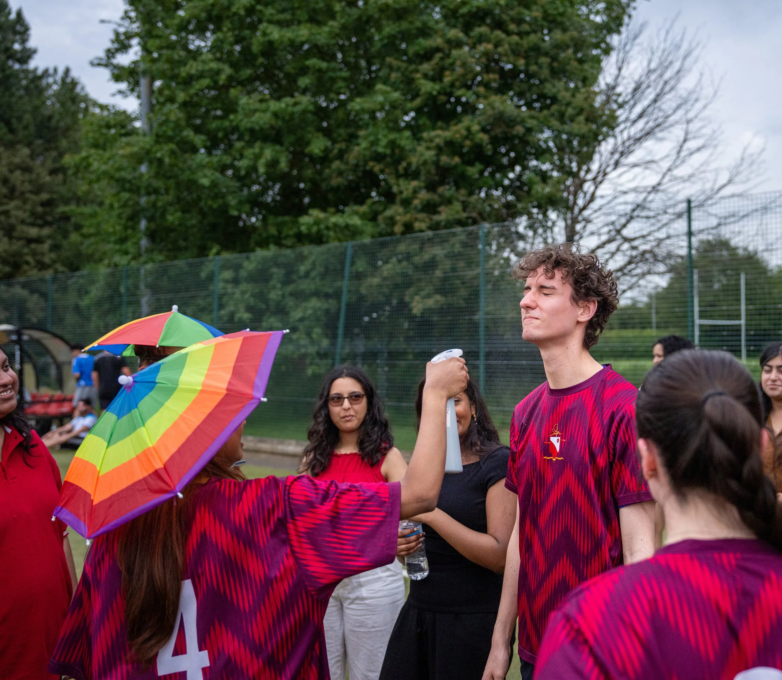 Group of people outdoors, some holding rainbow-colored umbrellas, engaging in conversation, with a large tree and a chain-link fence in the background.