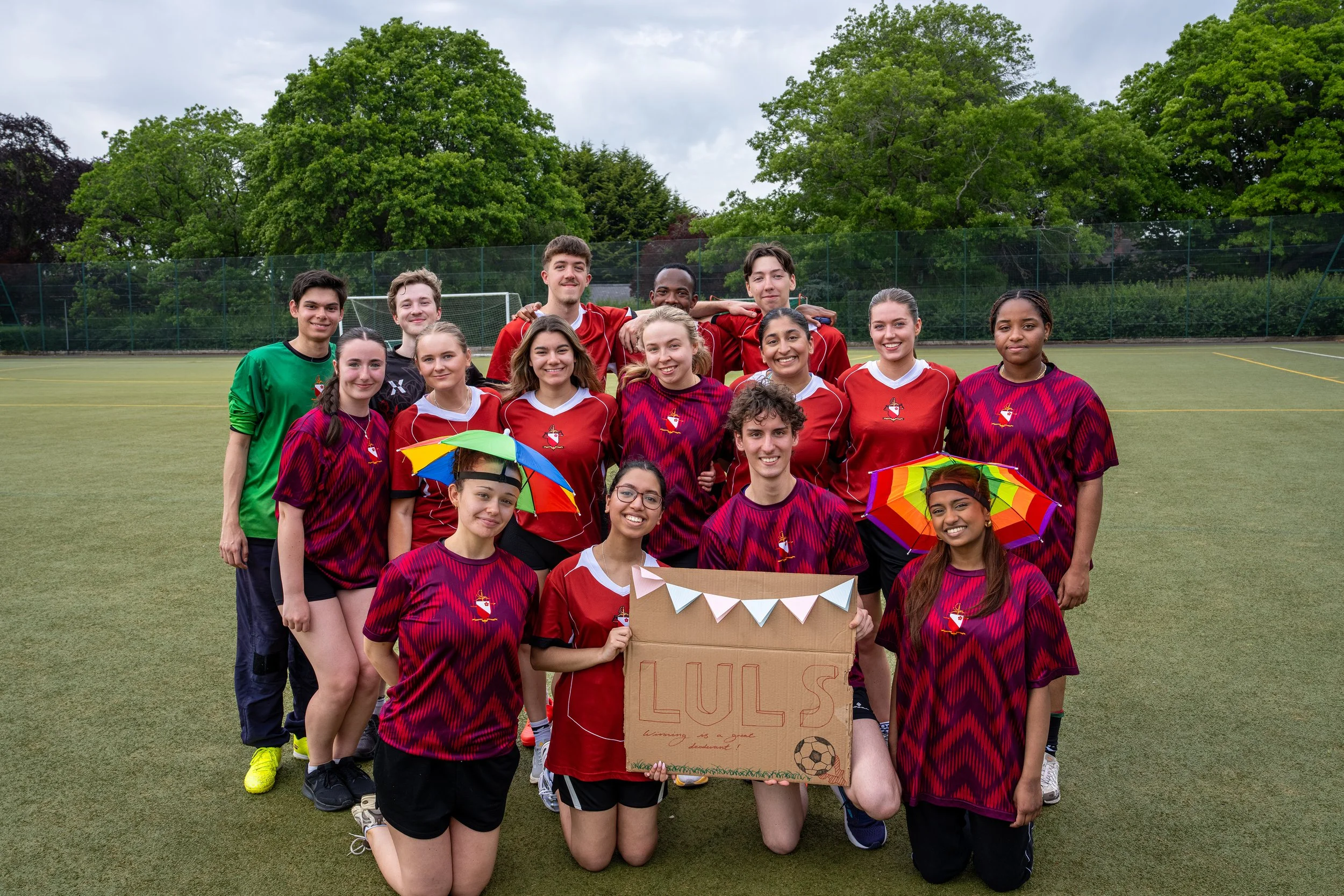 The Leicester University Law Society's charity football match team dressed in sports jerseys, holding a sign and colorful umbrellas, with trees and a cloudy sky in the background.