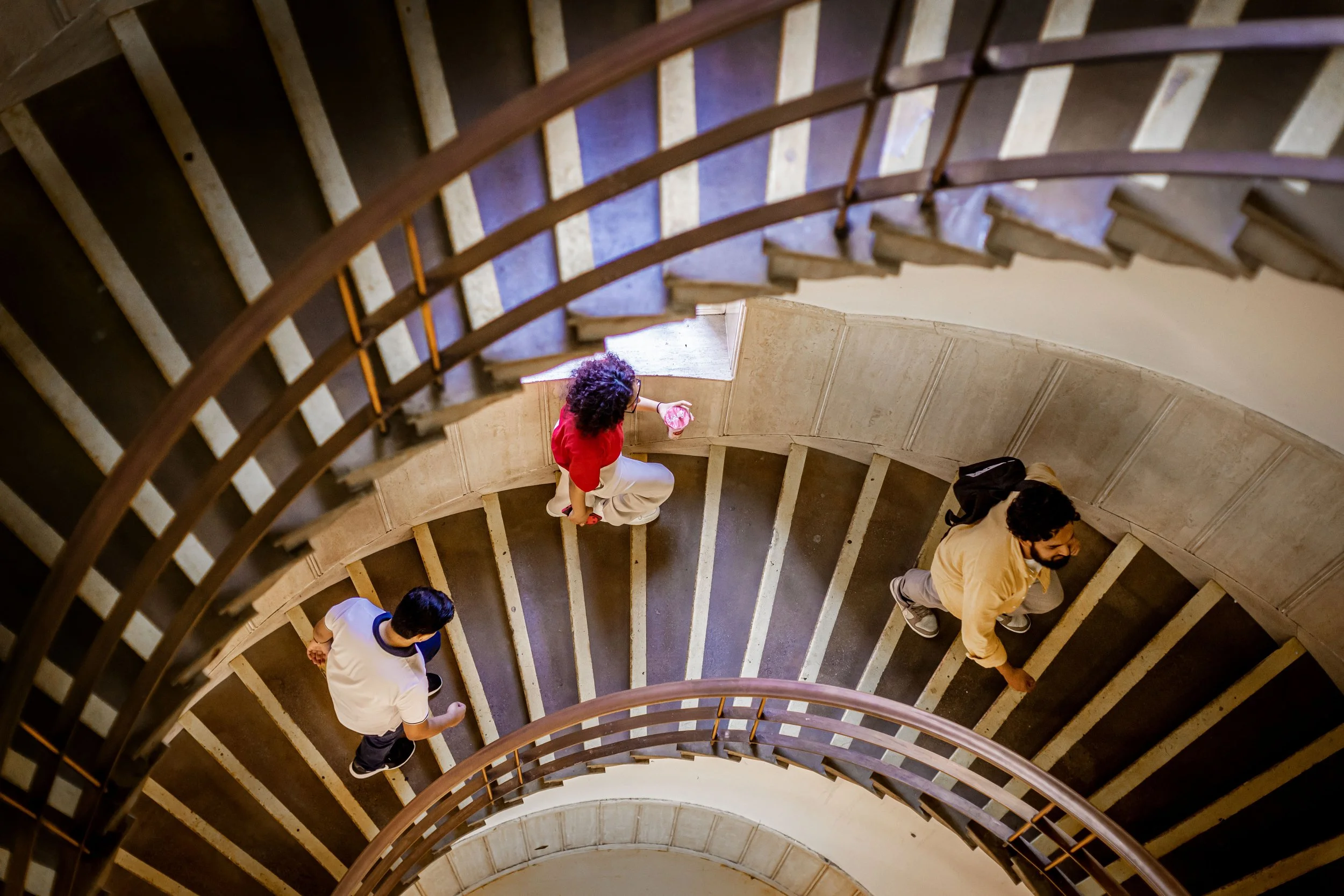 Overhead view of a spiral staircase with four people walking on it, two men and two women, with wooden steps and dark handrails.