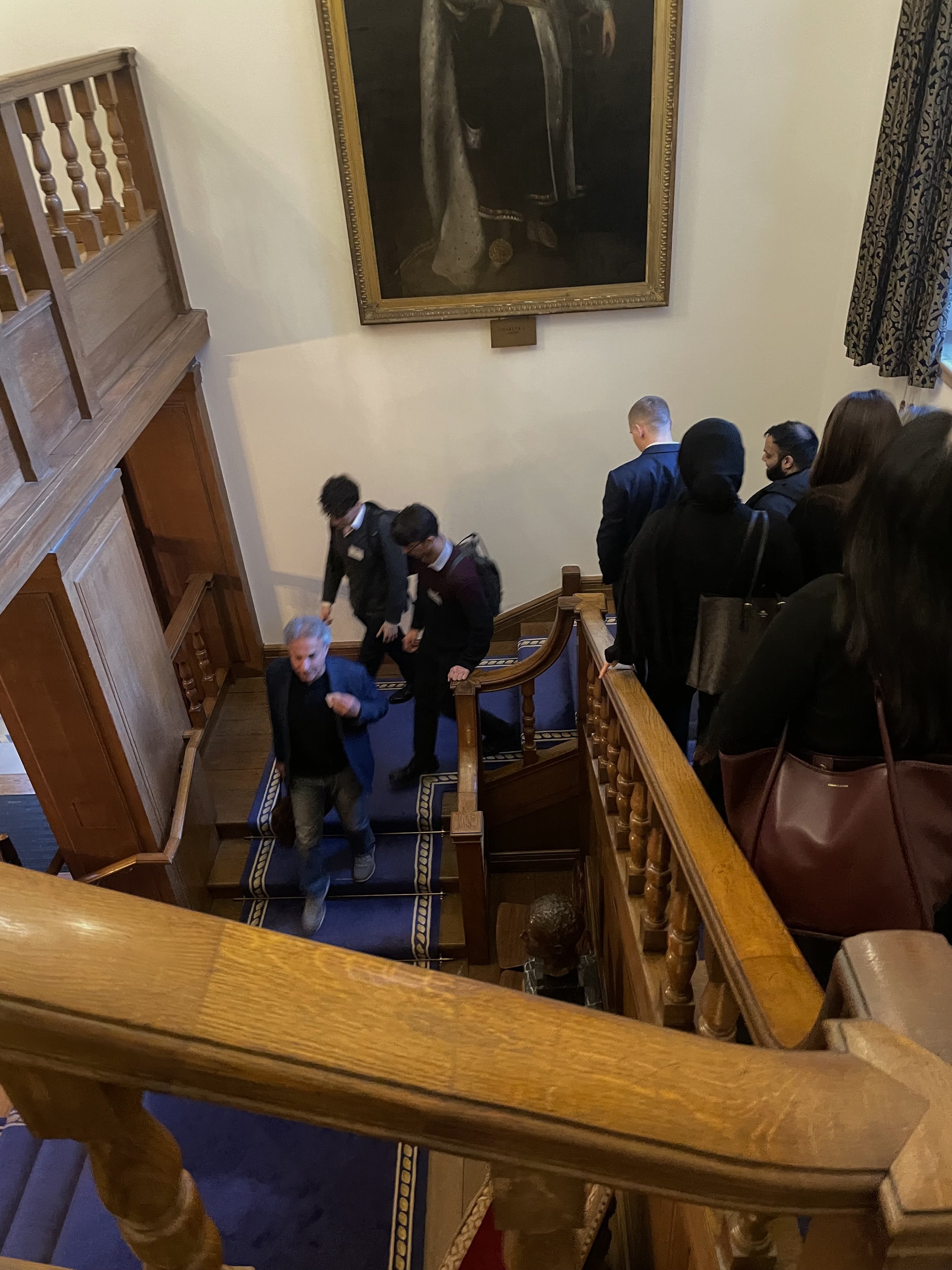 People walking up a wooden staircase inside a building with colonial-style decor. There is a large portrait painting on the wall at the top of the staircase and a curtain covering a window to the right.