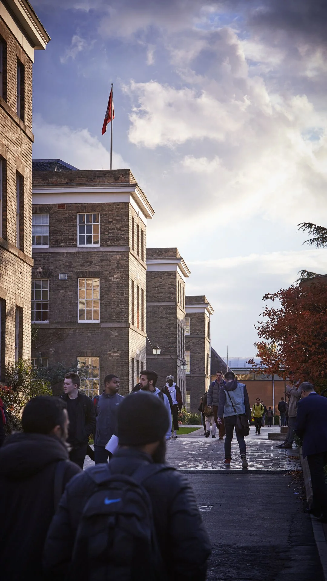 Students walking on the University of Leicester campus pathway with brick buildings and a flagpole with a red flag, trees with autumn leaves, under a partly cloudy sky.
