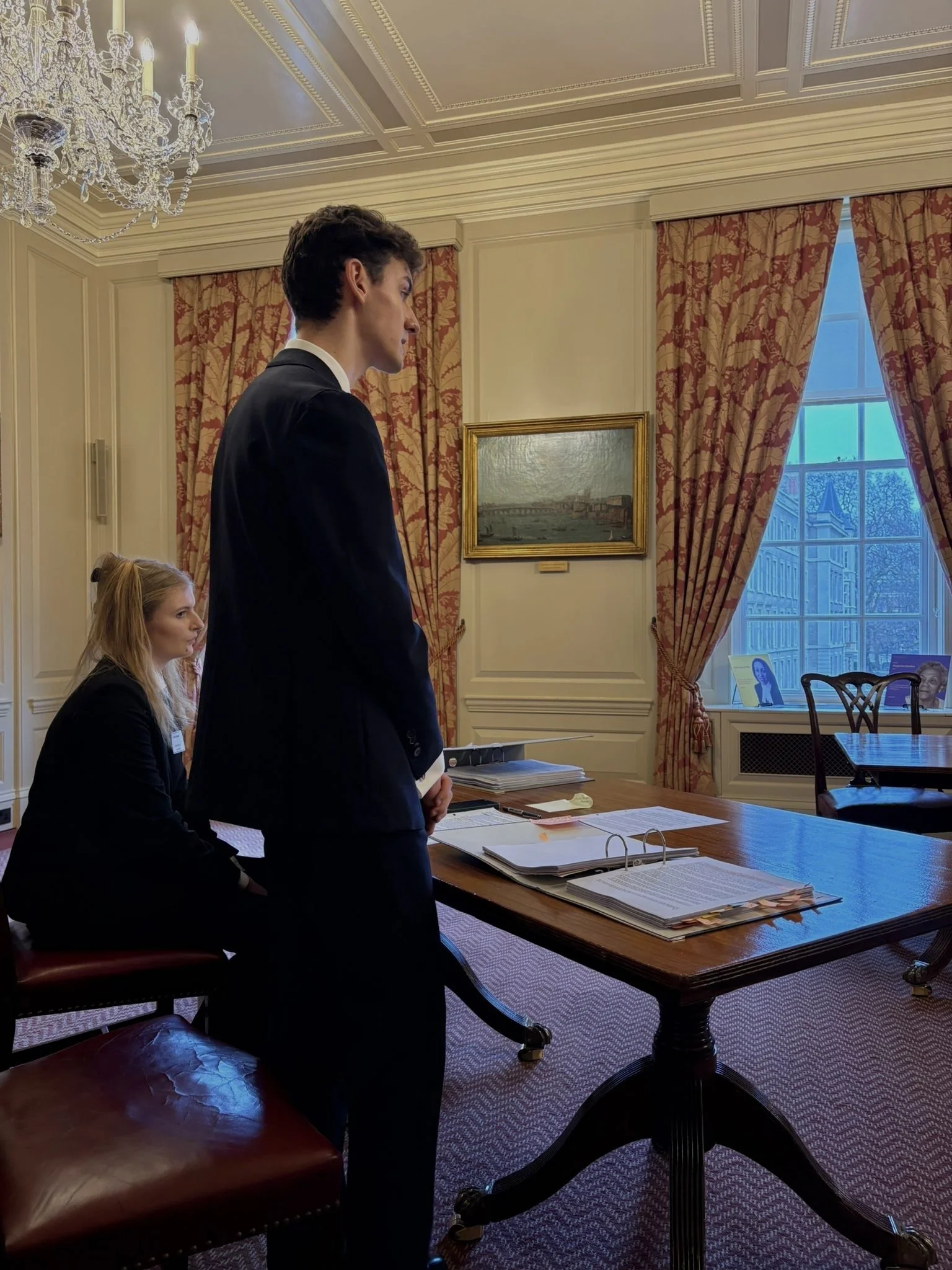 Thomas Wyatt and Sadie Worrall in formal business attire standing and sitting in a room with elegant decor, including a chandelier, paintings, and large windows with curtains.