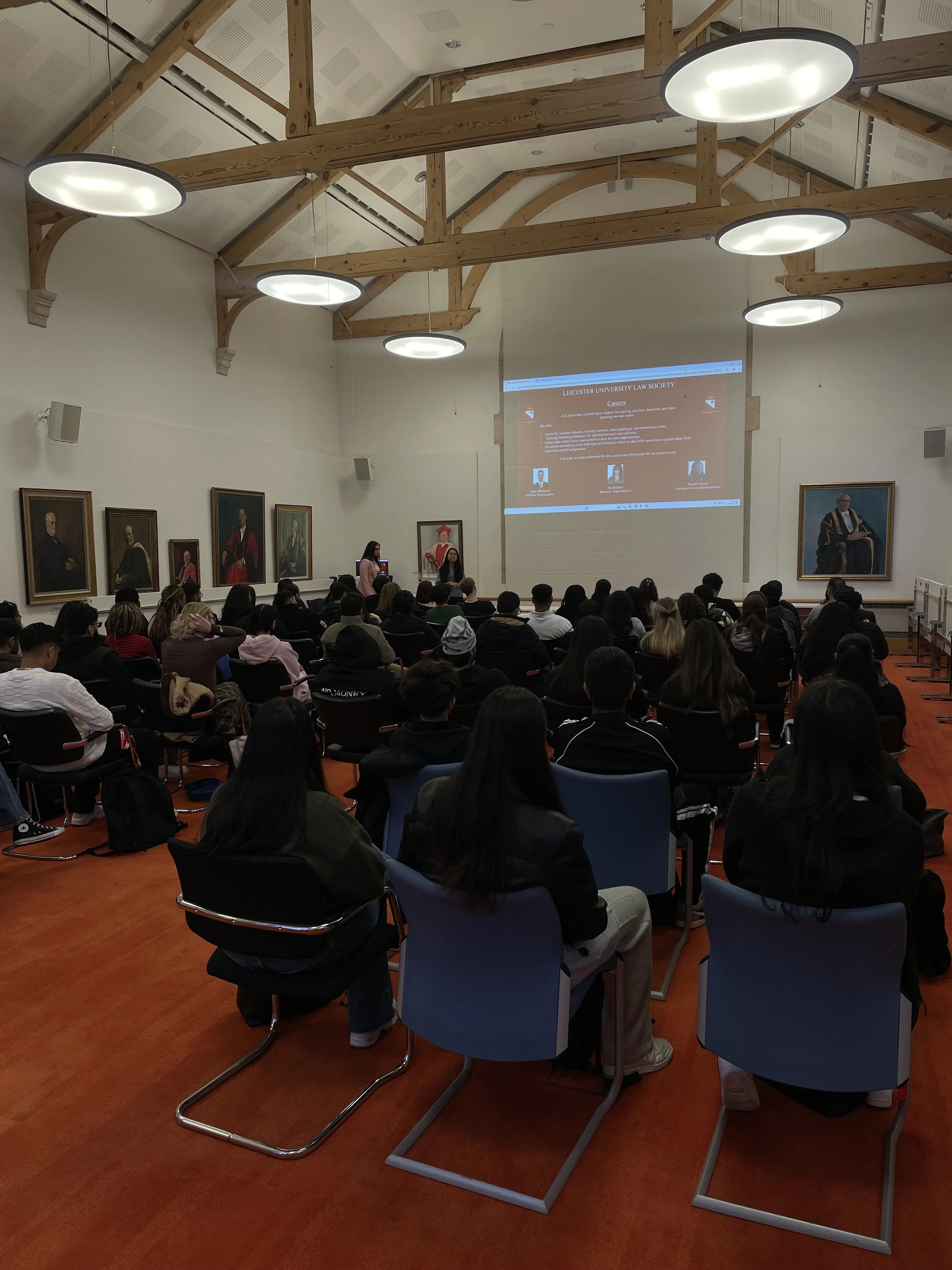 A lecture or presentation taking place in a large room with wooden beams and portraits on the walls. The audience is seated facing a large projection screen displaying a webpage from Leicester University Law Society. Two women are standing near the screen, one speaking and the other listening.