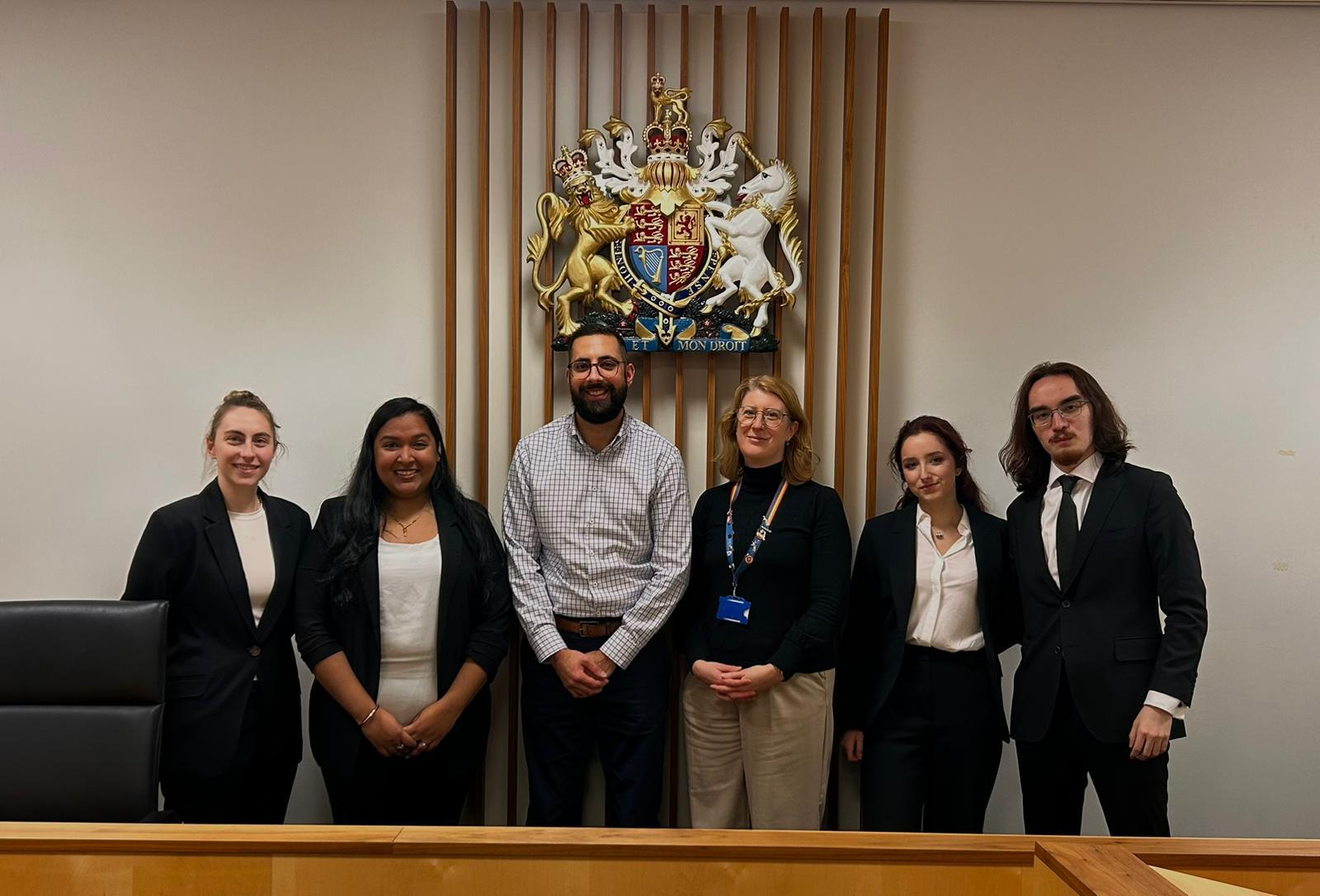 Group of six diverse people standing in front of a coat of arms in the University of Leicester's Moot Court, smiling at the camera.