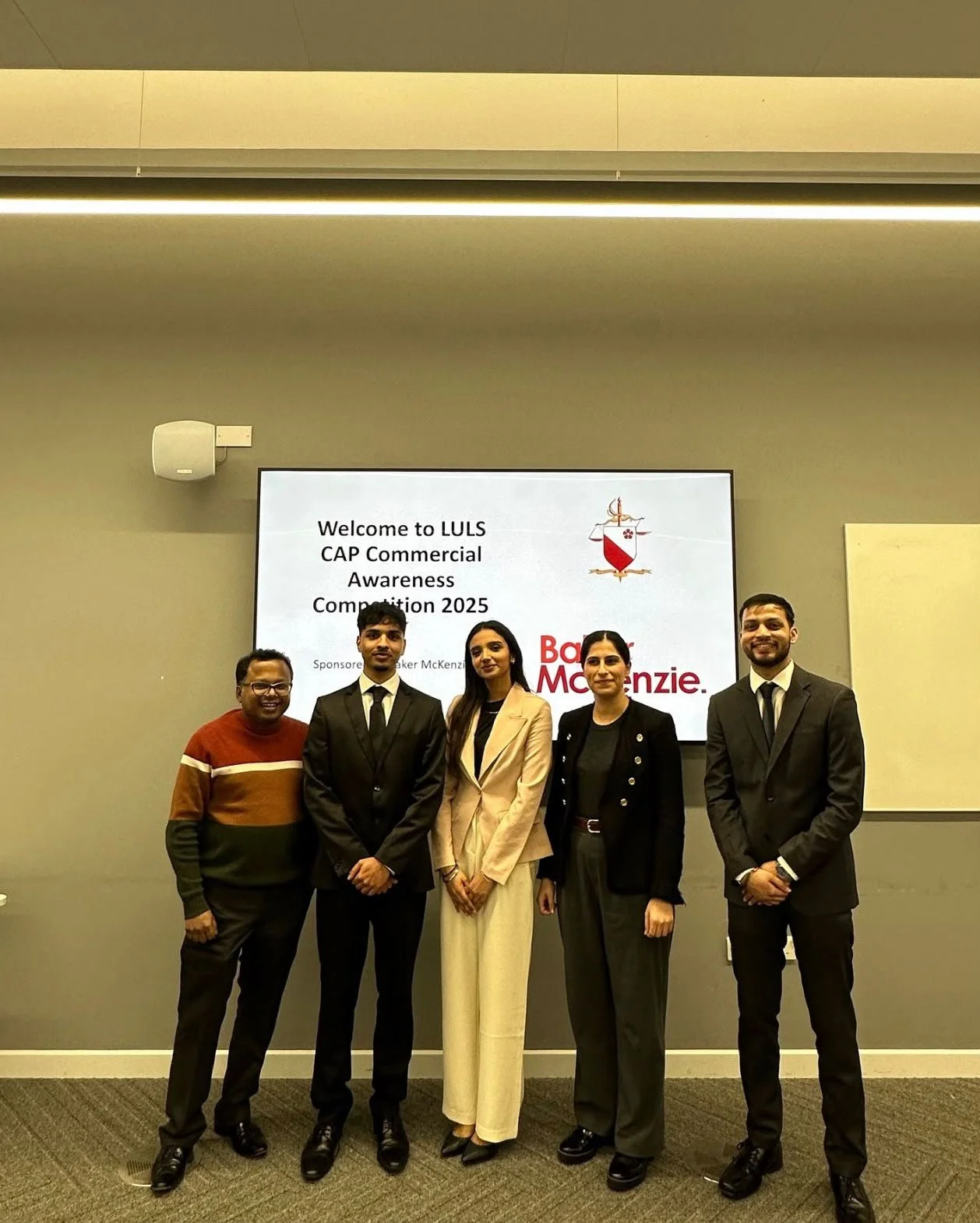 Group of five people in formal attire standing in front of a presentation screen at a corporate event. The screen displays welcome text for the LULS CAP Commercial Awareness Competition 2025 sponsored by Baker McKenzie. The event appears to be indoors with neutral-colored walls and ceiling lighting.