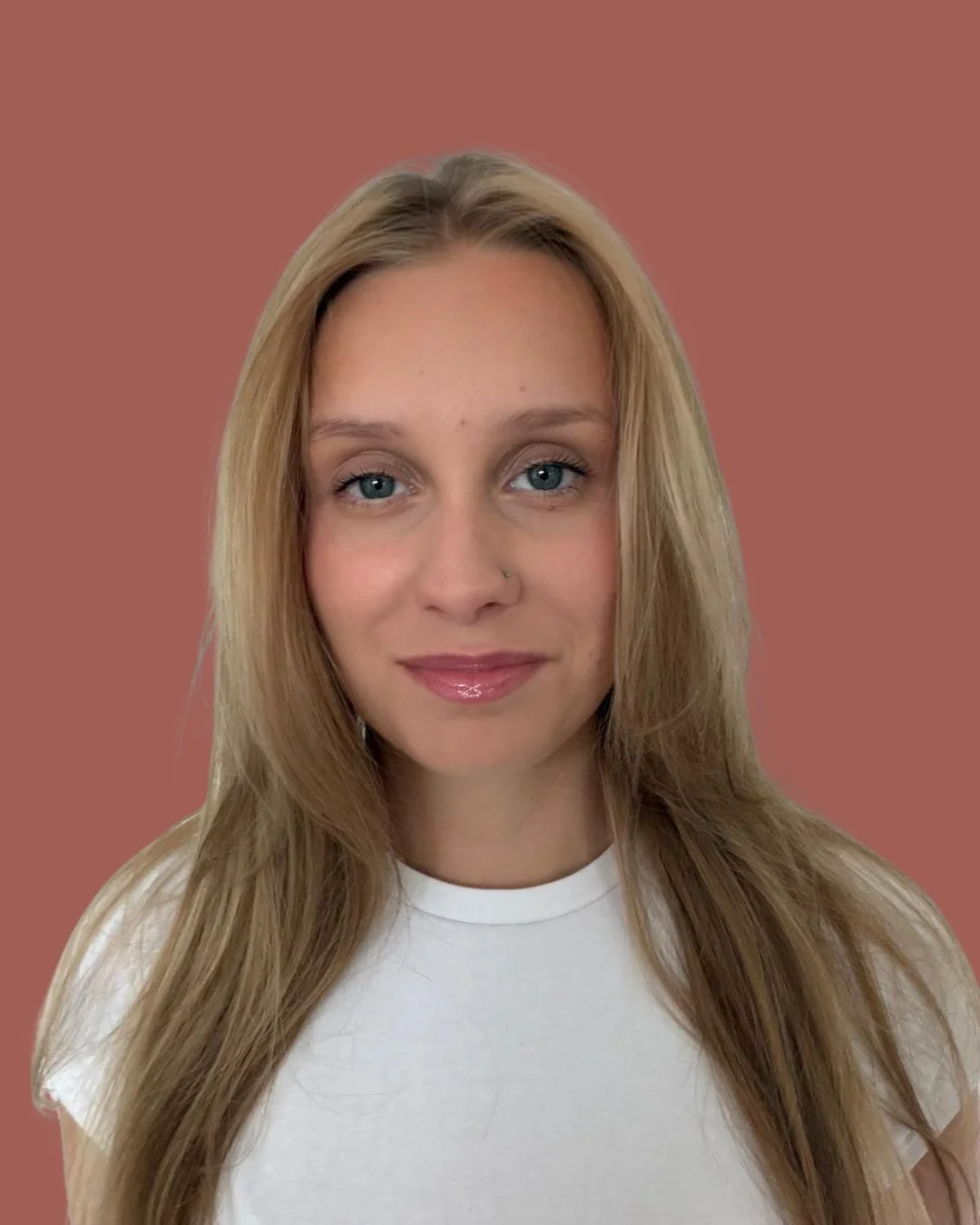 A woman with long blonde hair, blue eyes, wearing a white top, smiling slightly, with a solid muted pink background.