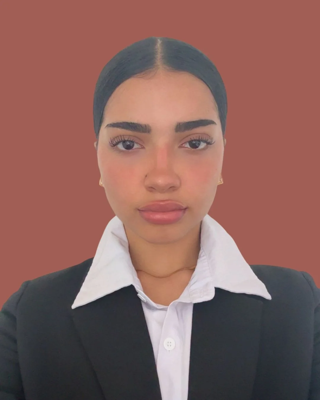 A young woman with dark hair pulled back, wearing a white collared shirt and black blazer, looking into the camera with a neutral expression, against a brown background.