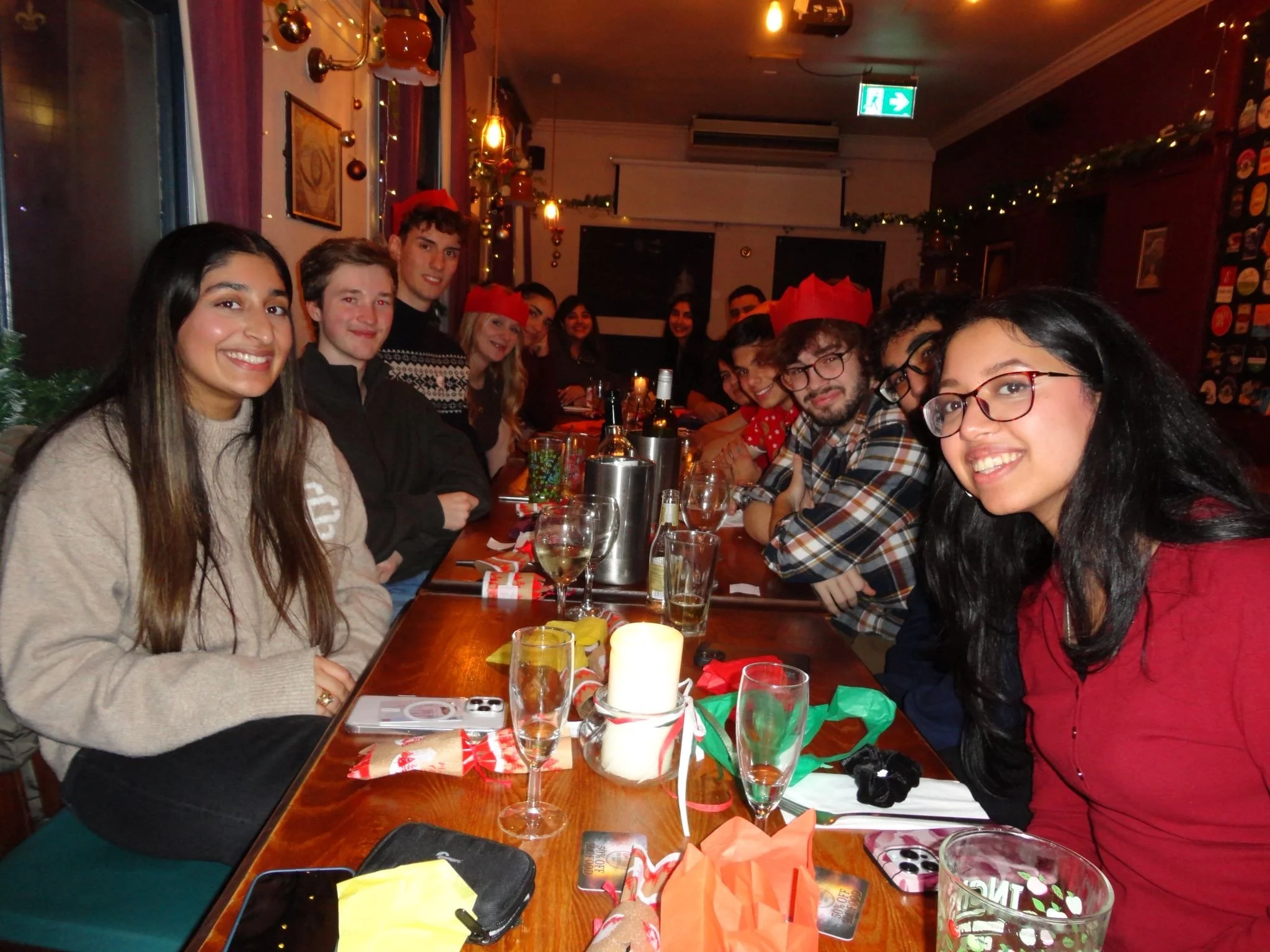 Group of people celebrating Christmas at a restaurant, sitting at a long table with Christmas decorations, wrapped gifts, wine glasses, and festive wrapped presents visible on the table.